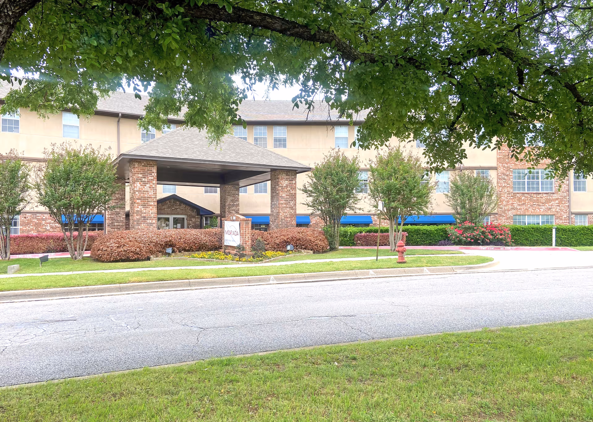 Exterior view of Morada Meadow Lakes senior living facility showing a three-story building with beige and brick facade, a covered entrance supported by brick pillars, landscaped bushes, trees, and a fire hydrant near the driveway.