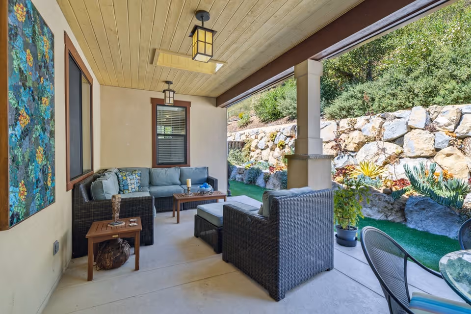 Covered outdoor patio area with wicker furniture including a sectional sofa, armchair, ottoman, and wooden side tables. The patio has a wooden ceiling with two hanging lantern-style lights and a skylight. A colorful floral painting is mounted on the wall. Beyond the patio is a rock retaining wall with various plants and greenery.