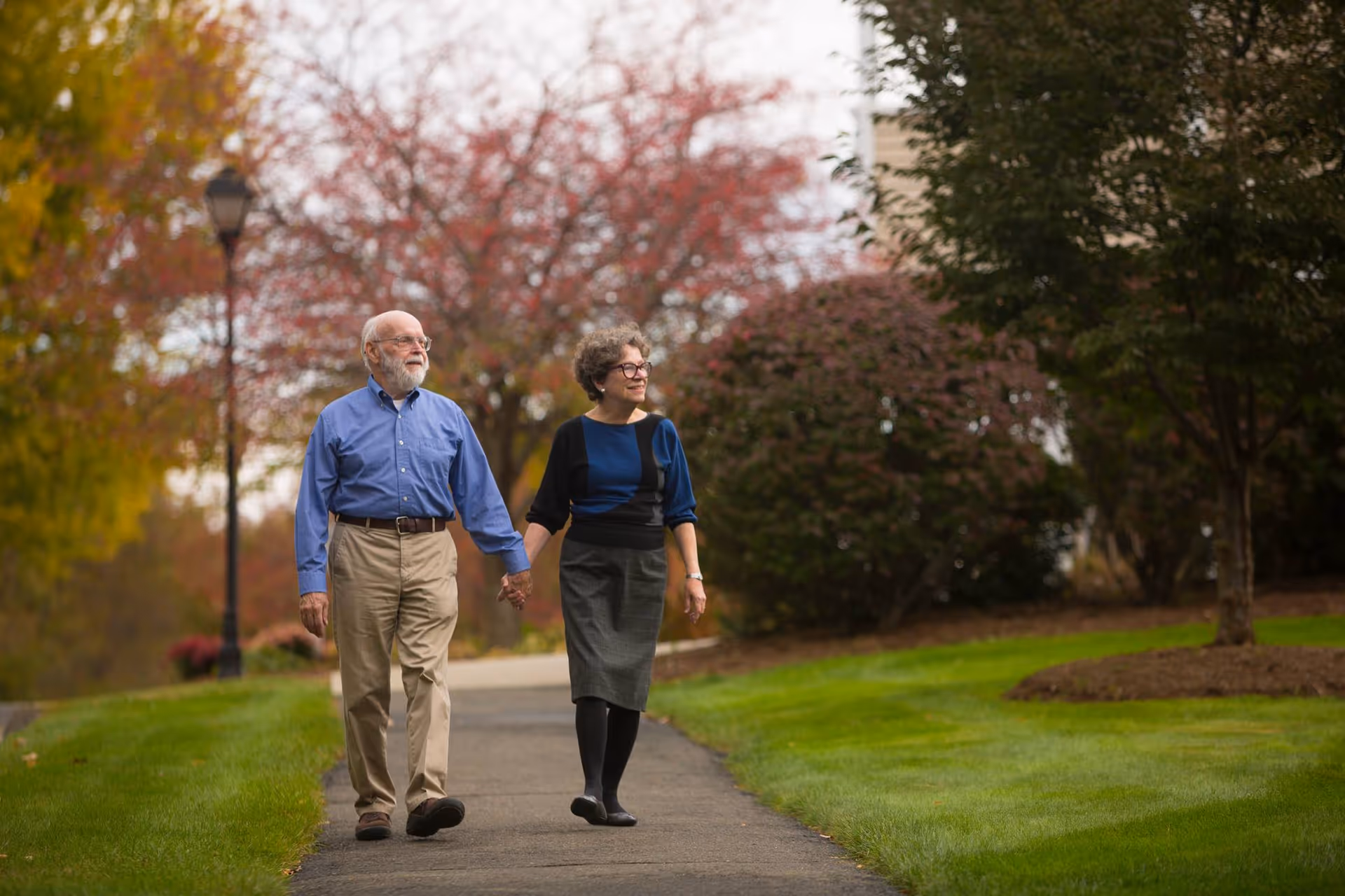 An elderly couple holding hands and walking along a paved path in a park-like setting with green grass, trees with autumn foliage, and a lamppost in the background.