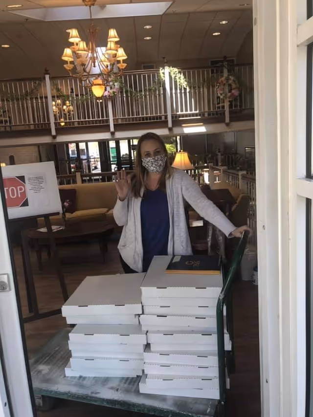 A woman wearing a face mask and casual clothing stands inside a room, waving at the camera. She is behind a cart stacked with multiple white pizza boxes. The room has a chandelier, a couch, a lamp, and a railing with decorative greenery.