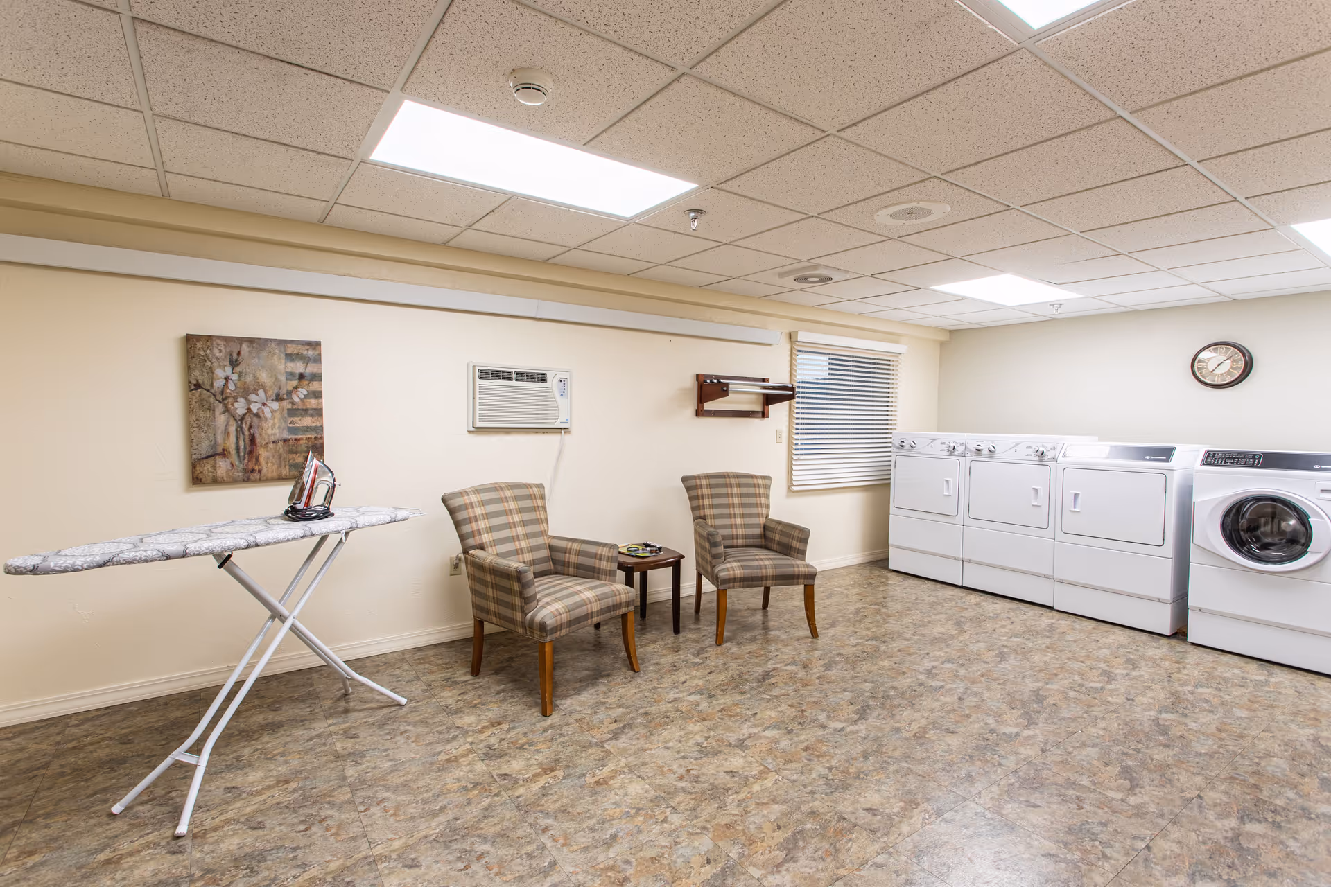 Laundry room with four white washing machines and dryers lined up against the wall. Two plaid upholstered chairs with wooden legs are placed next to a small wooden table. An ironing board with an iron on it is set up on the left side. The room has a beige tiled floor, cream-colored walls, a window with blinds, a wall-mounted air conditioner, and a clock on the wall.