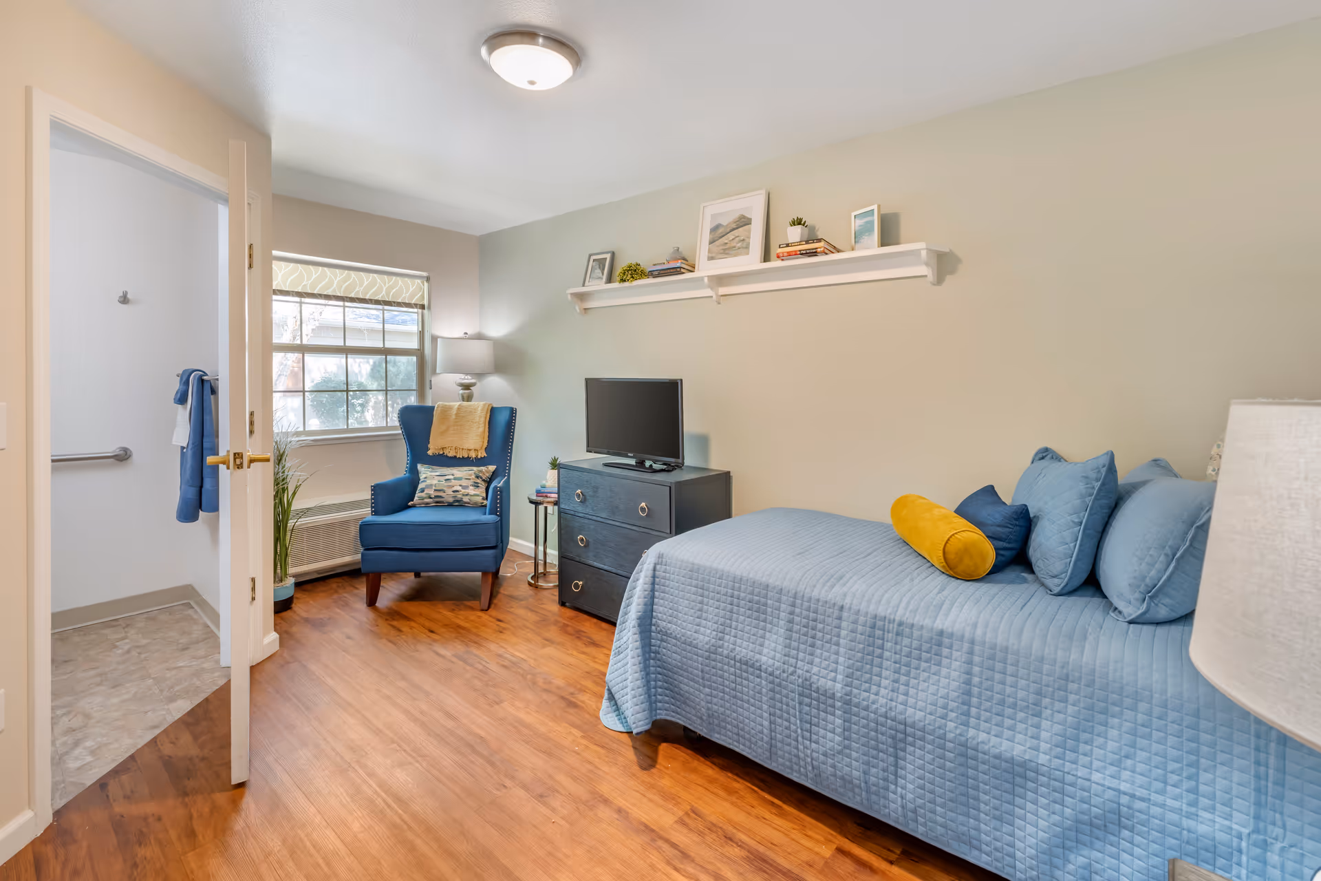 A cozy bedroom with a single bed covered in a light blue quilt and multiple pillows, including a yellow cylindrical one. There is a dark blue armchair with a patterned pillow and a yellow throw blanket near a window with a patterned roller shade. A small black dresser with a TV on top and a side table with books and a lamp are also visible. The room has wooden flooring and light-colored walls, with a shelf above the dresser holding framed pictures and small plants. An open door leads to a bathroom with tiled flooring and a towel hanging on a grab bar.