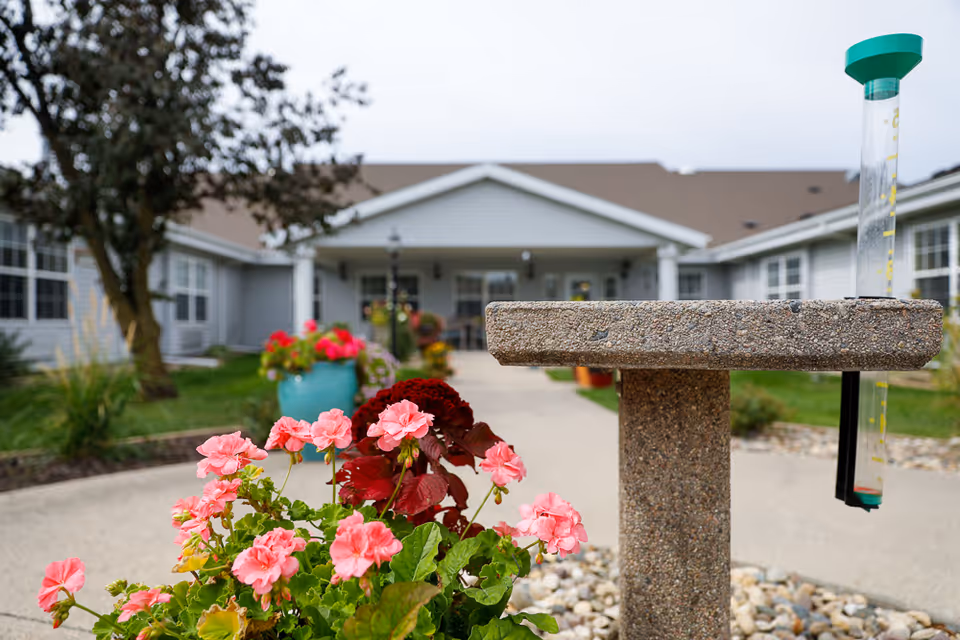 Outdoor view of a senior living facility with a concrete birdbath or rain gauge in the foreground, surrounded by pink flowers and greenery. The building is visible in the background with a covered entrance and multiple windows.