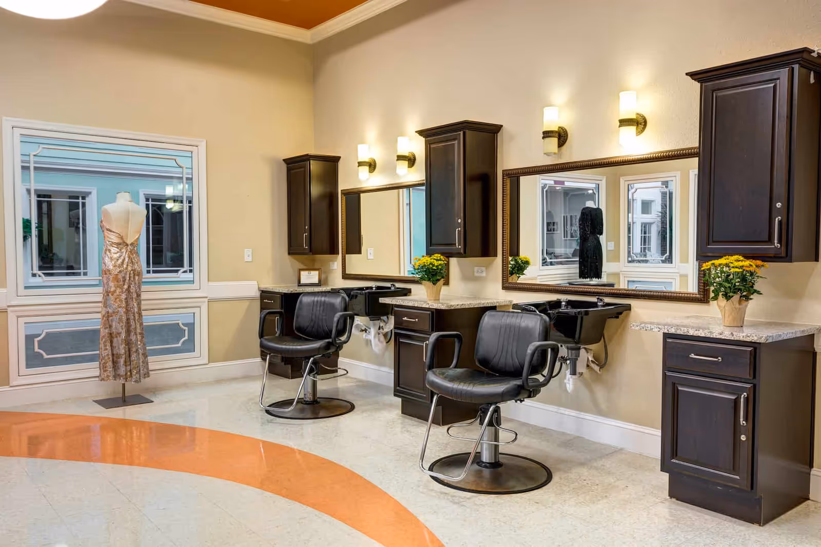 Interior view of a salon area with two black salon chairs in front of large mirrors mounted on a beige wall. Each station has dark wood cabinets and a small sink. There are wall-mounted lights above the mirrors and a mannequin dressed in a gold and white gown near a window.