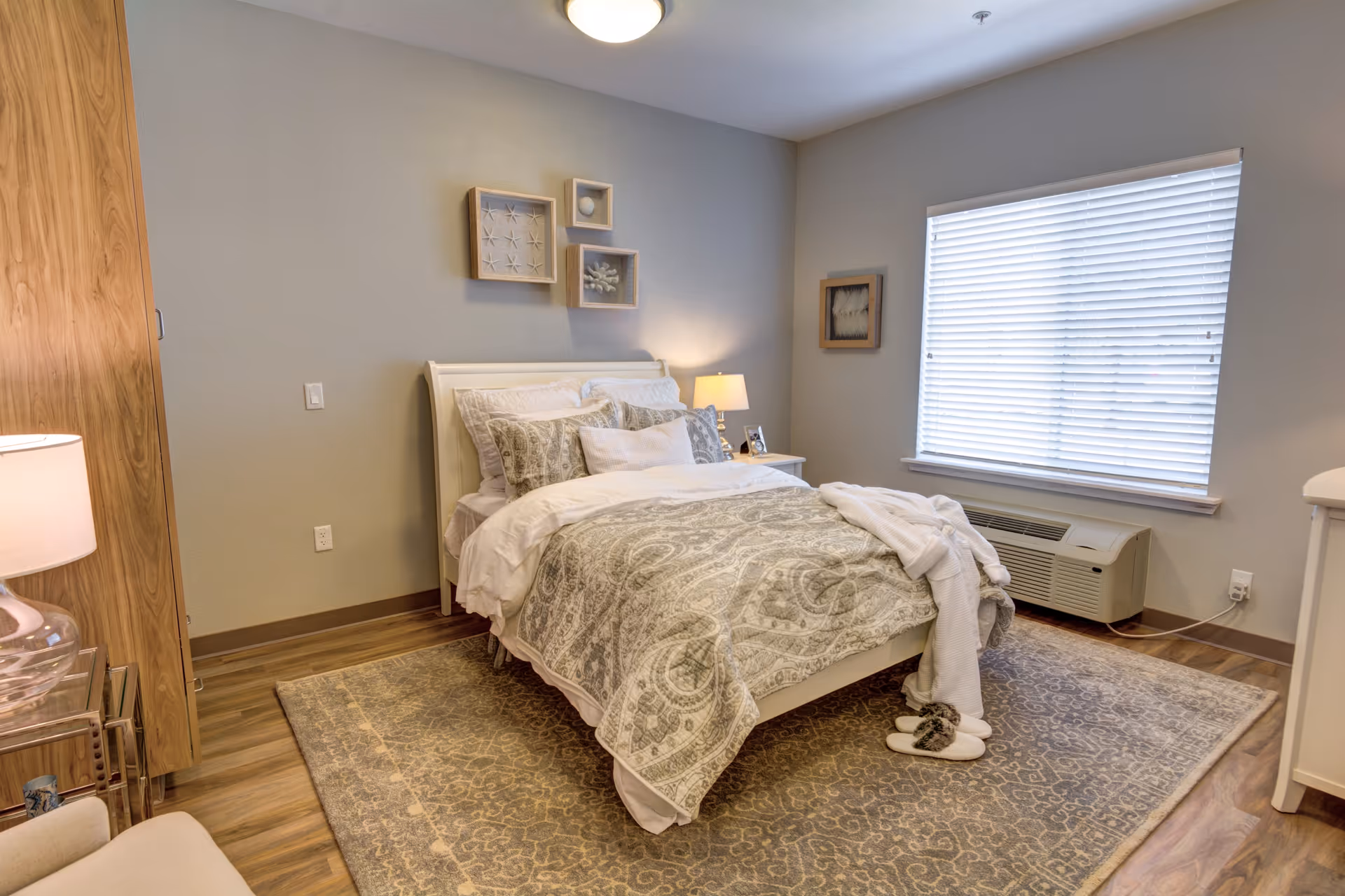 A cozy bedroom with a neatly made bed featuring patterned bedding and multiple pillows. The room has a large window with blinds, a nightstand with a lamp and picture frame, a wooden wardrobe, a patterned area rug, and a pair of slippers on the floor near the bed.