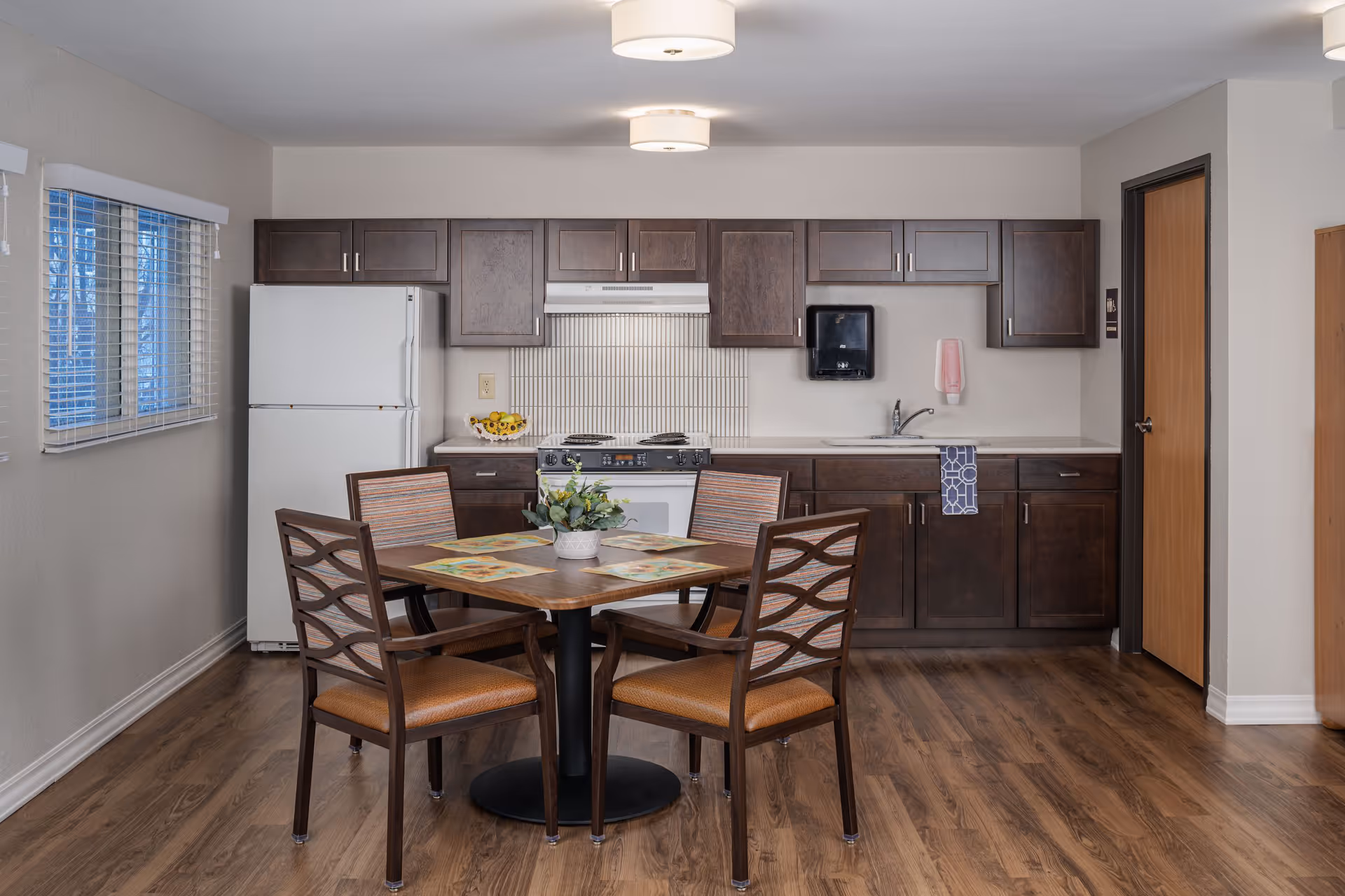 A clean and well-lit kitchen area with dark wood cabinets, a white refrigerator, stove, and sink. In the center, there is a wooden dining table with four chairs, each with a cushioned seat. The floor is wood, and there is a window with blinds on the left wall. Two ceiling lights illuminate the space.
