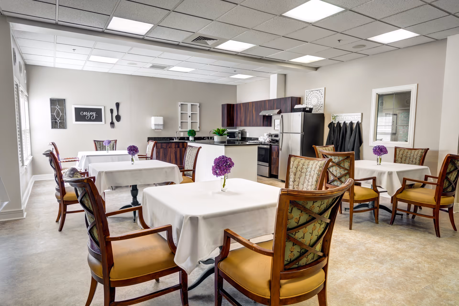 A bright dining room with several tables covered in white tablecloths, each adorned with a small vase of purple flowers. The room features wooden chairs with patterned upholstery and a kitchen area in the background with dark wood cabinets, a stainless steel refrigerator, stove, and microwave. The walls are decorated with framed art and utensils, and there is a window with blinds on the left side.