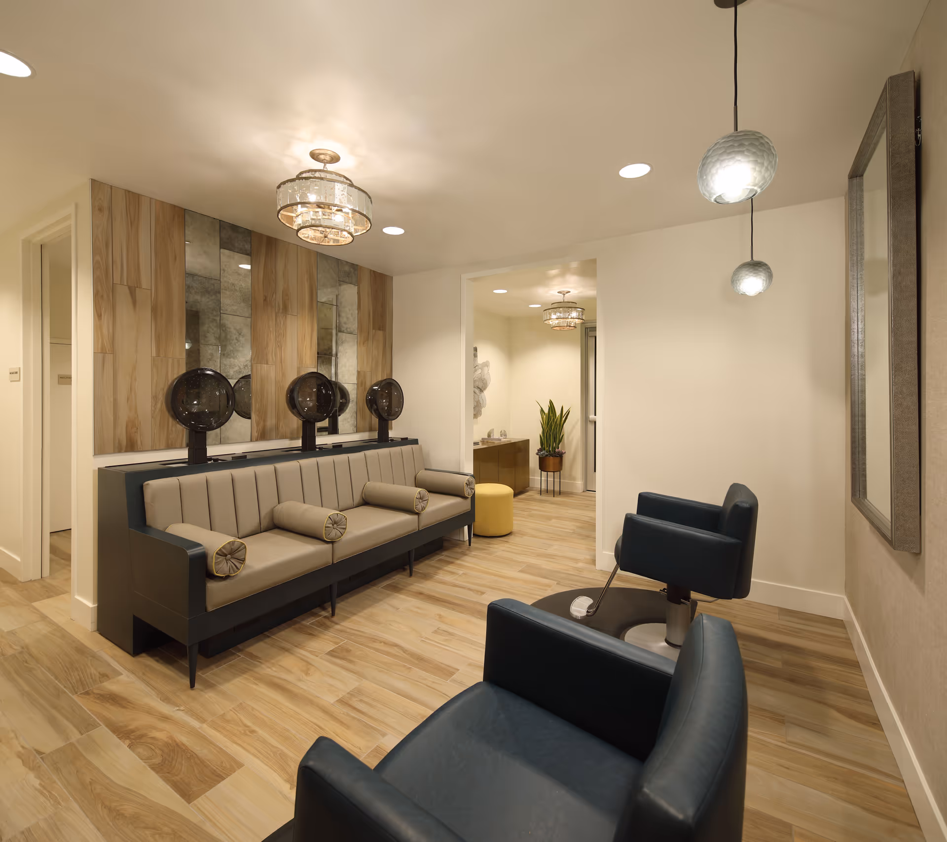 A modern salon area in a senior living facility featuring a long cushioned bench with three vintage-style hair dryers mounted above it, two black salon chairs, wooden flooring, a large wall mirror, and soft overhead lighting fixtures.
