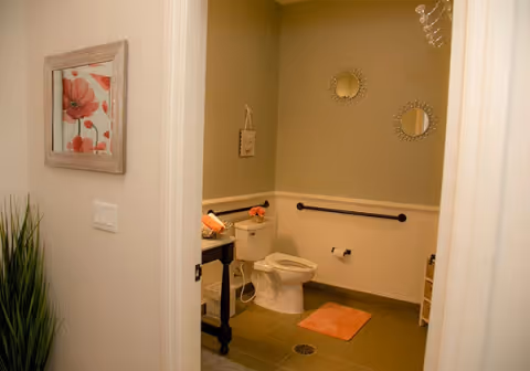 View into a bathroom with a white toilet, a small black table with towels and flowers, two decorative round mirrors on the wall, a peach-colored bath mat on the floor, and a grab bar next to the toilet. The walls are painted beige and white, and there is a framed floral picture on the outside wall near the door.