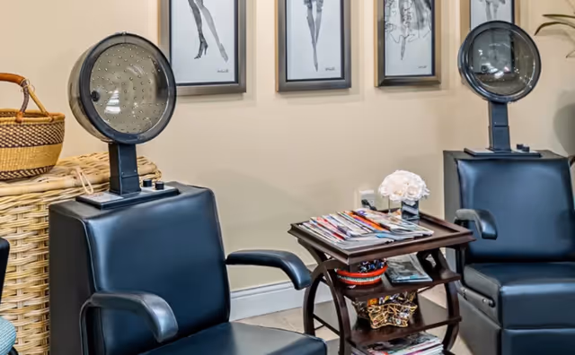 Interior view of a senior living facility hair salon area with two black salon chairs equipped with hair dryers. Between the chairs is a small wooden table holding magazines and a small flower arrangement. The wall behind features framed black and white artwork.