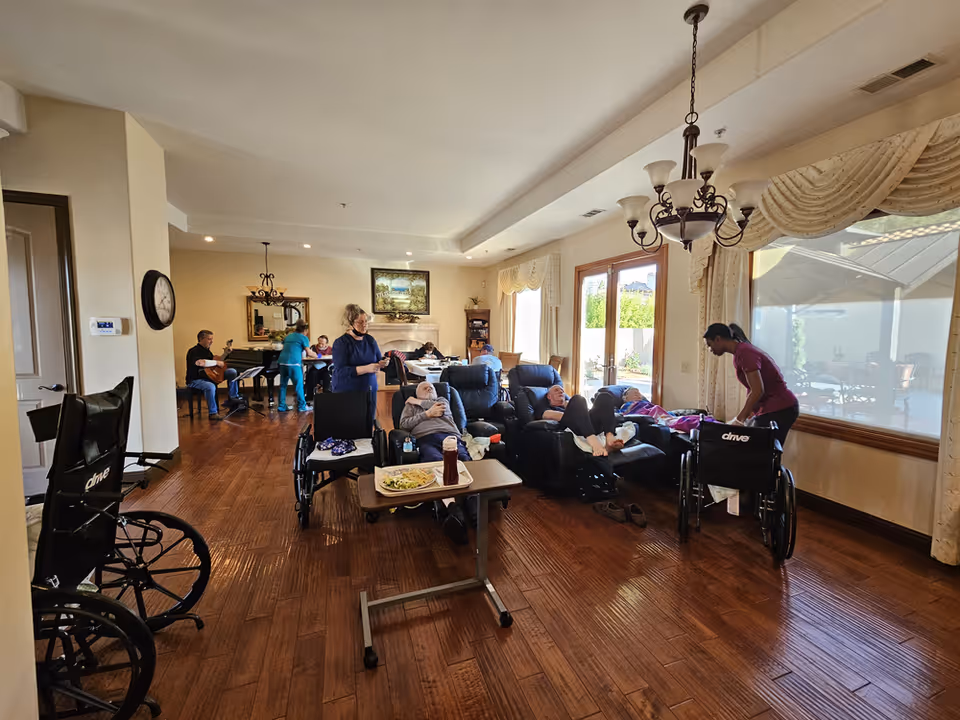 A spacious living room in a senior care home with wooden flooring and large windows letting in natural light. Several elderly residents are seated in recliners and wheelchairs, some being assisted by caregivers. In the background, a man is playing a guitar near a piano, and other people are engaged in activities around a table. The room is warmly decorated with curtains, a chandelier, and a painting above a fireplace.