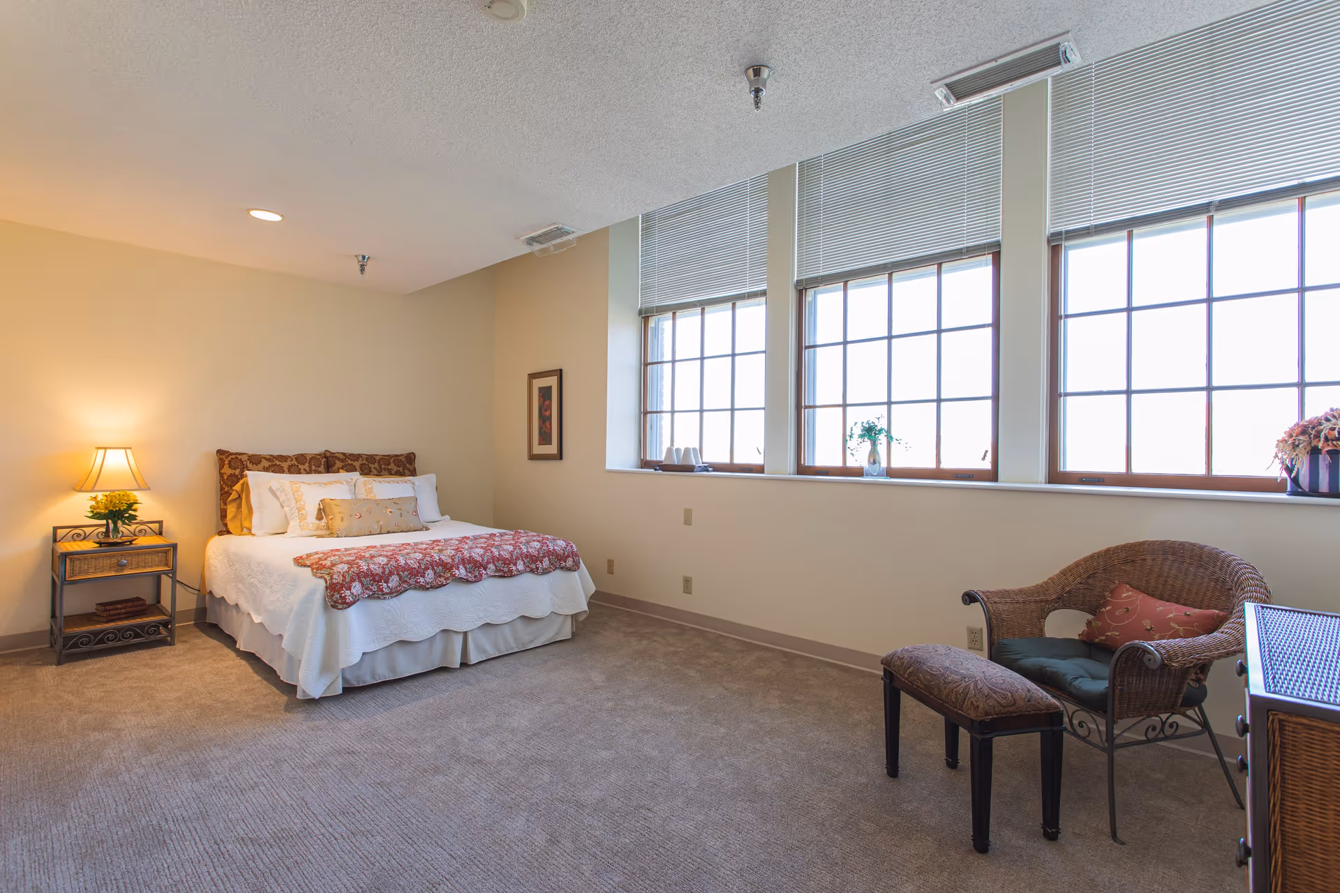 A cozy bedroom in Old Main Village featuring a bed with white and floral bedding, a bedside table with a lamp and flowers, a wicker chair with cushions, a small bench, and large windows with blinds letting in natural light.