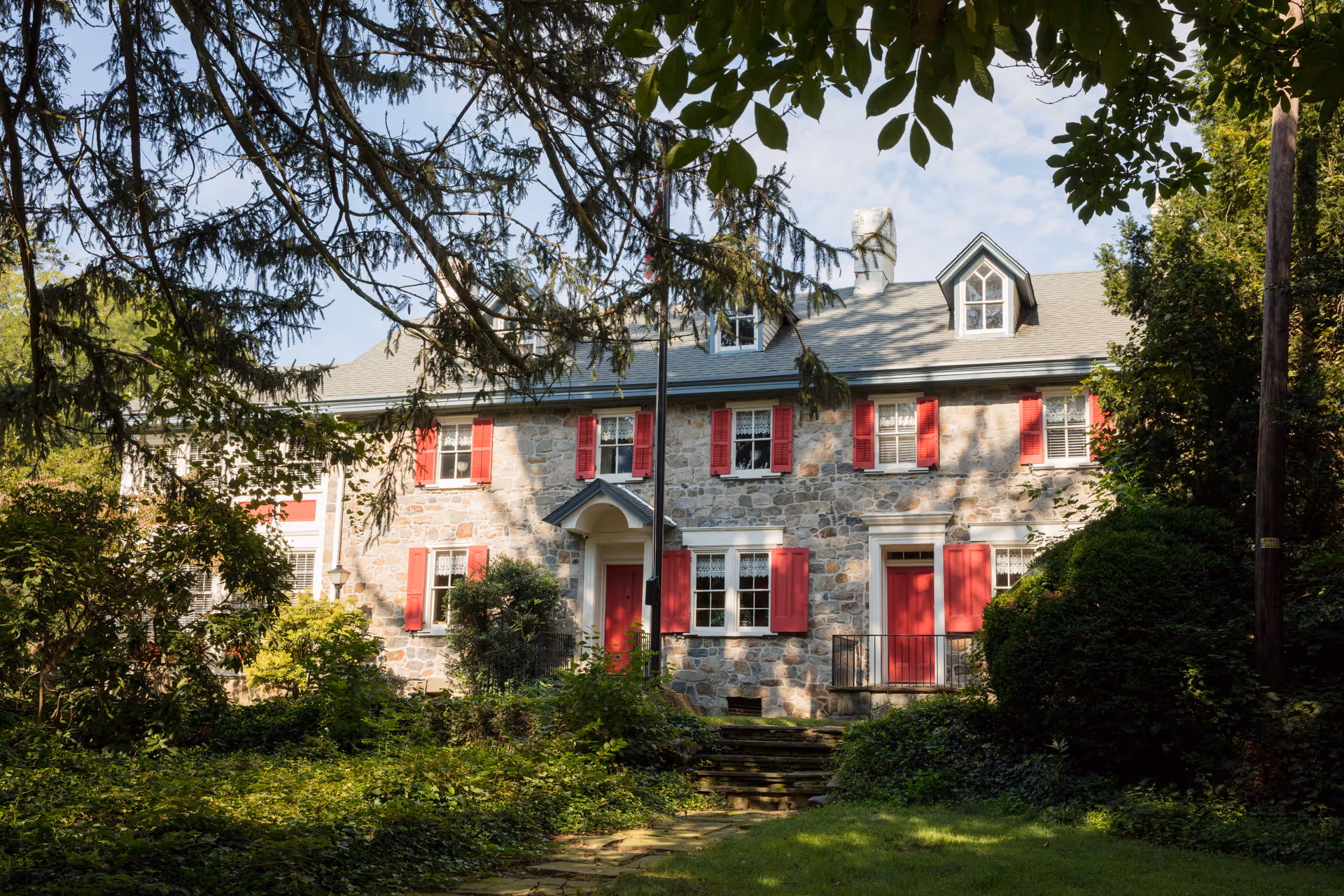 Stone building with red shutters and doors surrounded by trees and greenery under a partly cloudy sky.