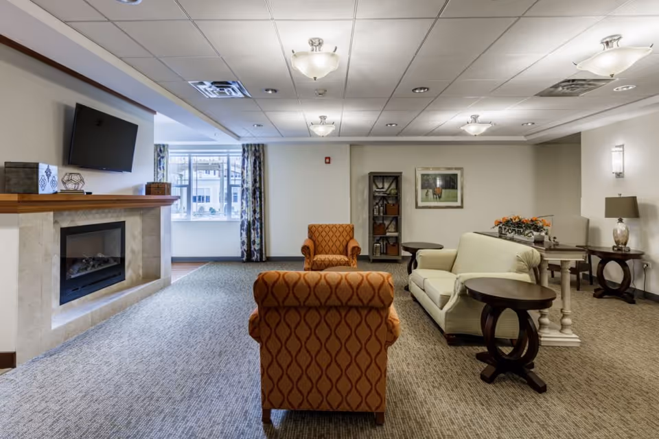 A cozy living room area in a senior living facility featuring a fireplace with a mounted TV above it, two patterned orange armchairs, a light-colored sofa, several side tables, a bookshelf, and a window with patterned curtains allowing natural light in.