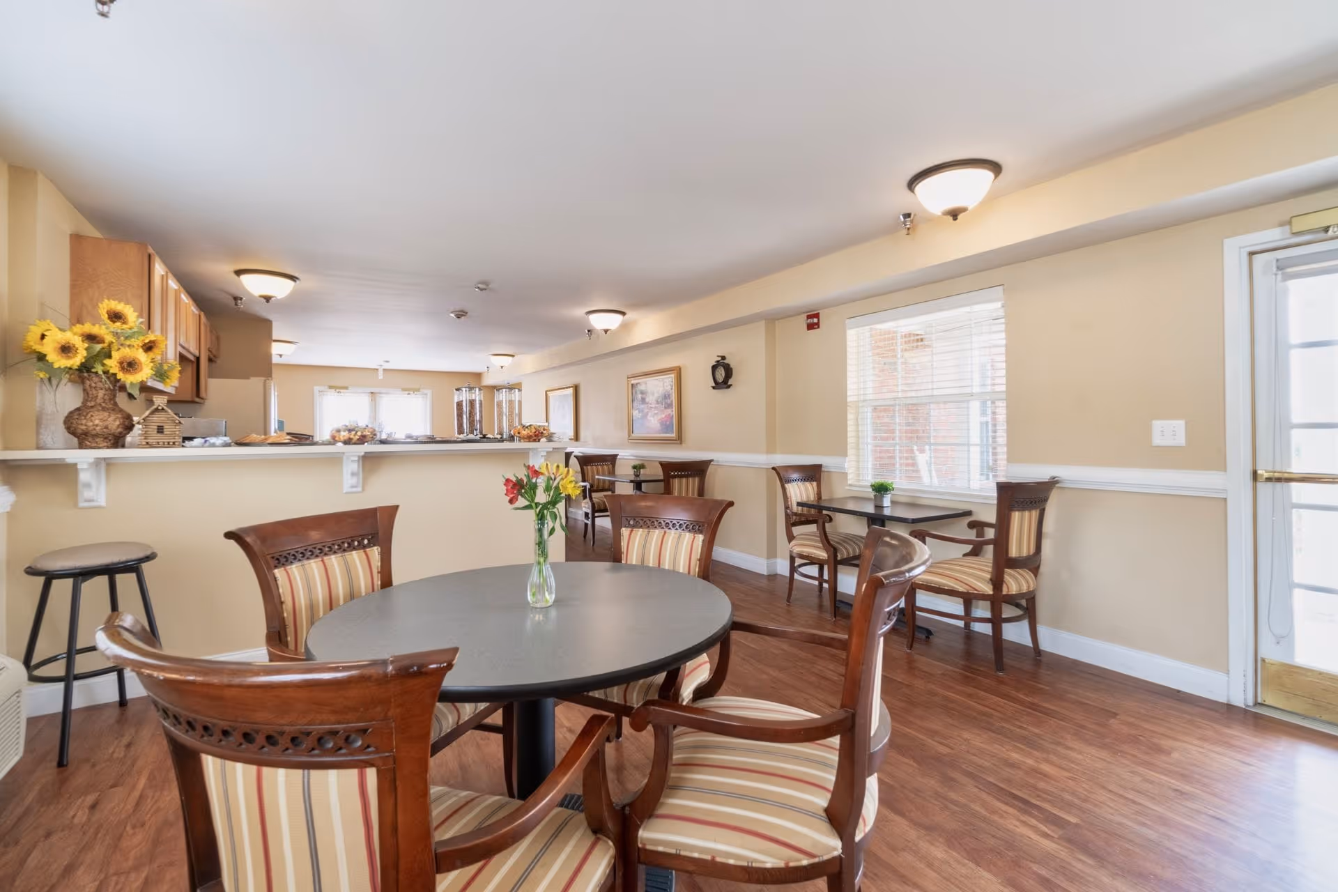 Bright communal dining area with round tables and striped chairs, a serving counter with sunflowers, and windows letting in natural light.