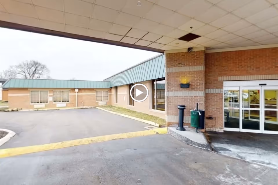 Entrance area of a senior living facility with a covered drop-off zone, brick walls, glass sliding doors, and a paved driveway. There are windows on the building and some outdoor fixtures like a trash can and a cigarette disposal stand.