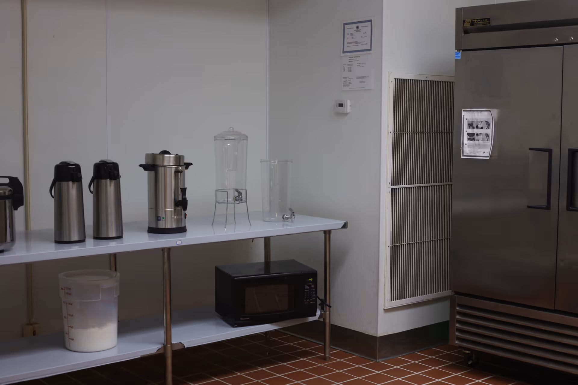 Interior view of a kitchen area with a white table holding two stainless steel airpots, a large coffee urn, a clear beverage dispenser, and a clear plastic container. Below the table is a black microwave and a large plastic container with white powder. To the right is a large stainless steel refrigerator with papers attached to it. The floor is tiled with reddish-brown tiles.