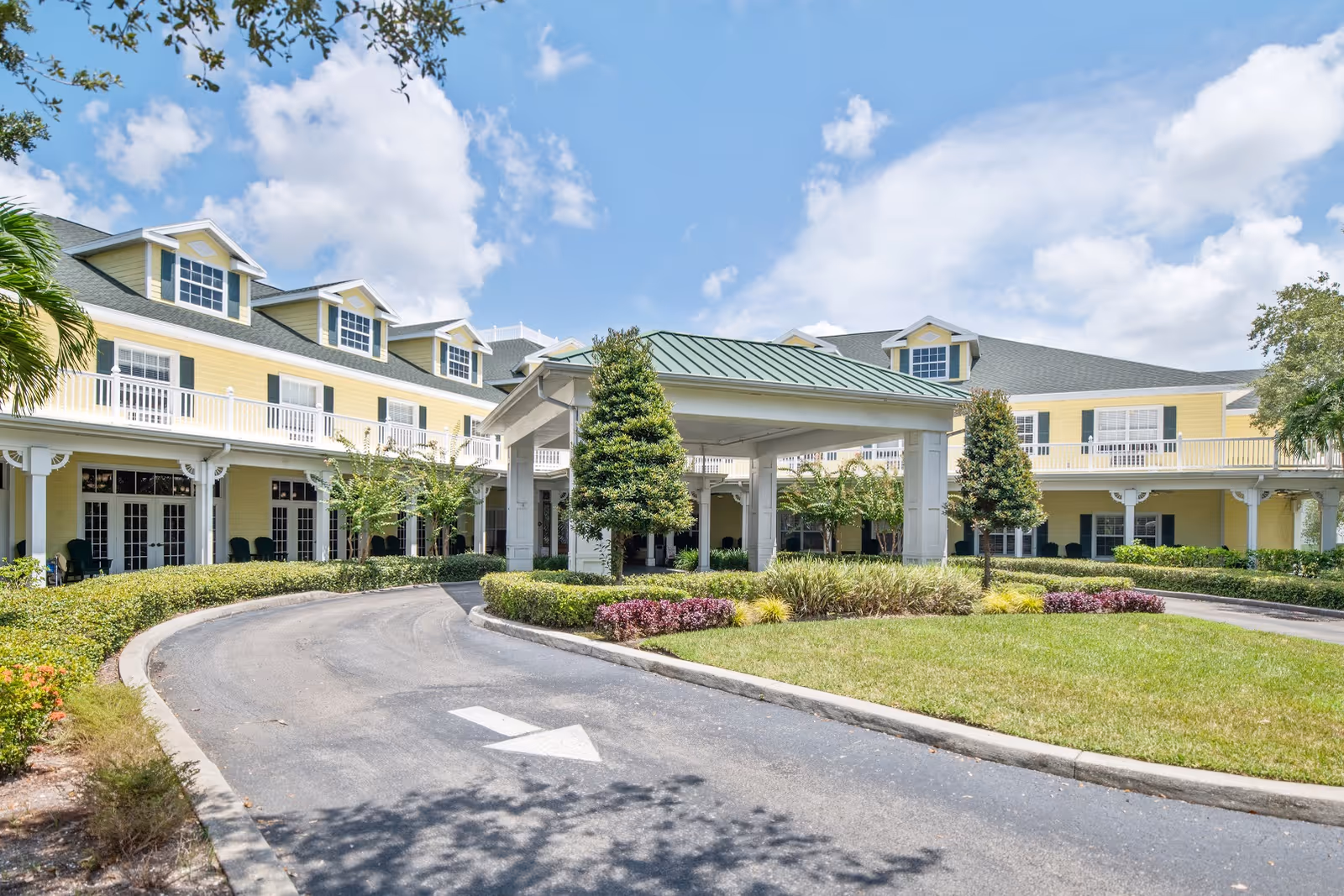 Exterior view of a yellow two-story senior living facility with white trim and a green roof over the entrance driveway. The building has multiple windows and dormers, surrounded by well-maintained landscaping including trees, bushes, and grass under a partly cloudy blue sky.