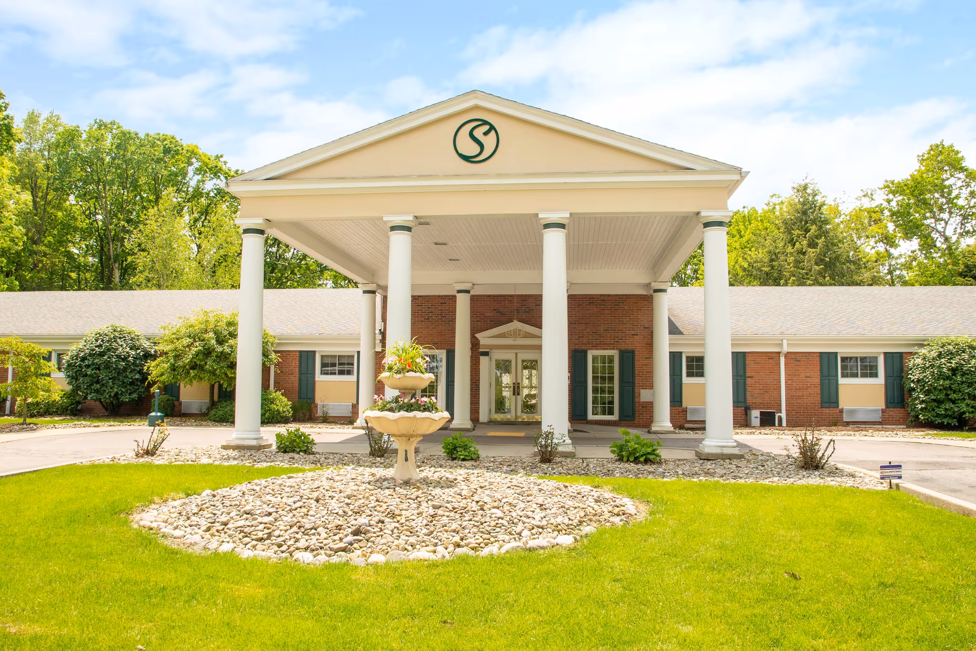 Front exterior view of Shelbourne Personal Care facility featuring a covered entrance with white columns, a circular stone garden with a tiered planter in the center, green lawn, and surrounding trees under a partly cloudy sky.