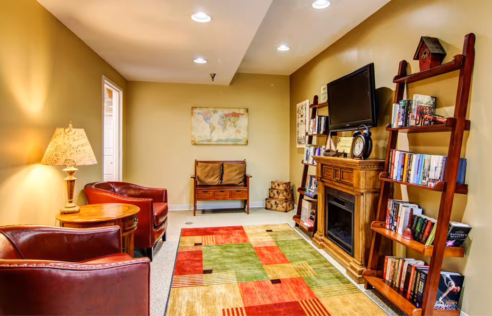 A cozy senior living community lounge with two red leather armchairs, a round wooden side table with a decorative lamp, a wooden bench with cushions, a colorful area rug, a wooden electric fireplace with a clock and decorative items on top, two wooden ladder-style bookshelves filled with books, and a wall-mounted flat-screen TV. The walls are painted beige and there is a world map artwork hanging on the far wall.