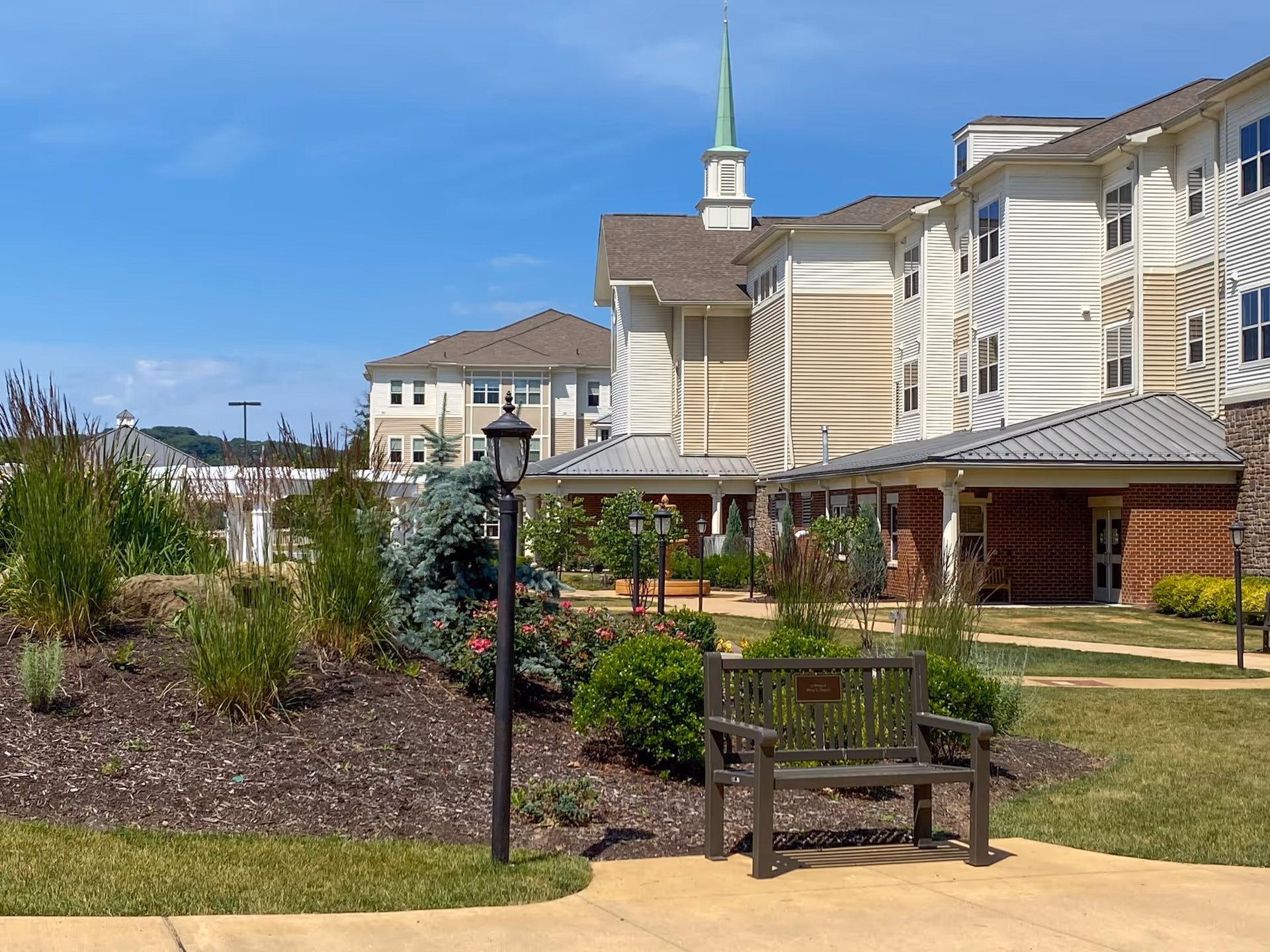 Outdoor courtyard of a senior living facility with landscaped beds, a bench and lampposts in front of a multi-story building with a small steeple.
