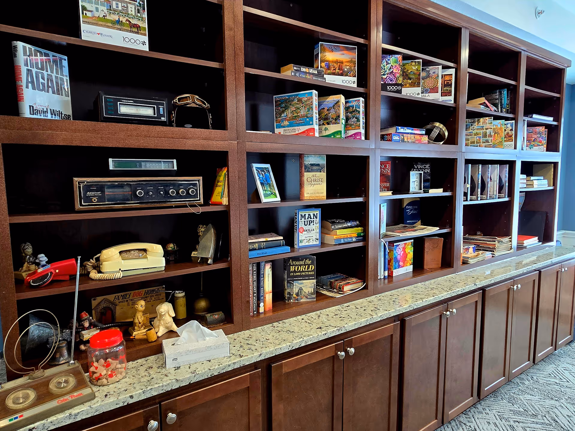 A large wooden bookshelf with multiple compartments filled with books, puzzles, vintage radios, a rotary telephone, and various decorative items. The bookshelf has a granite countertop and cabinets below. The setting appears to be a cozy common area or lounge in a senior living facility.