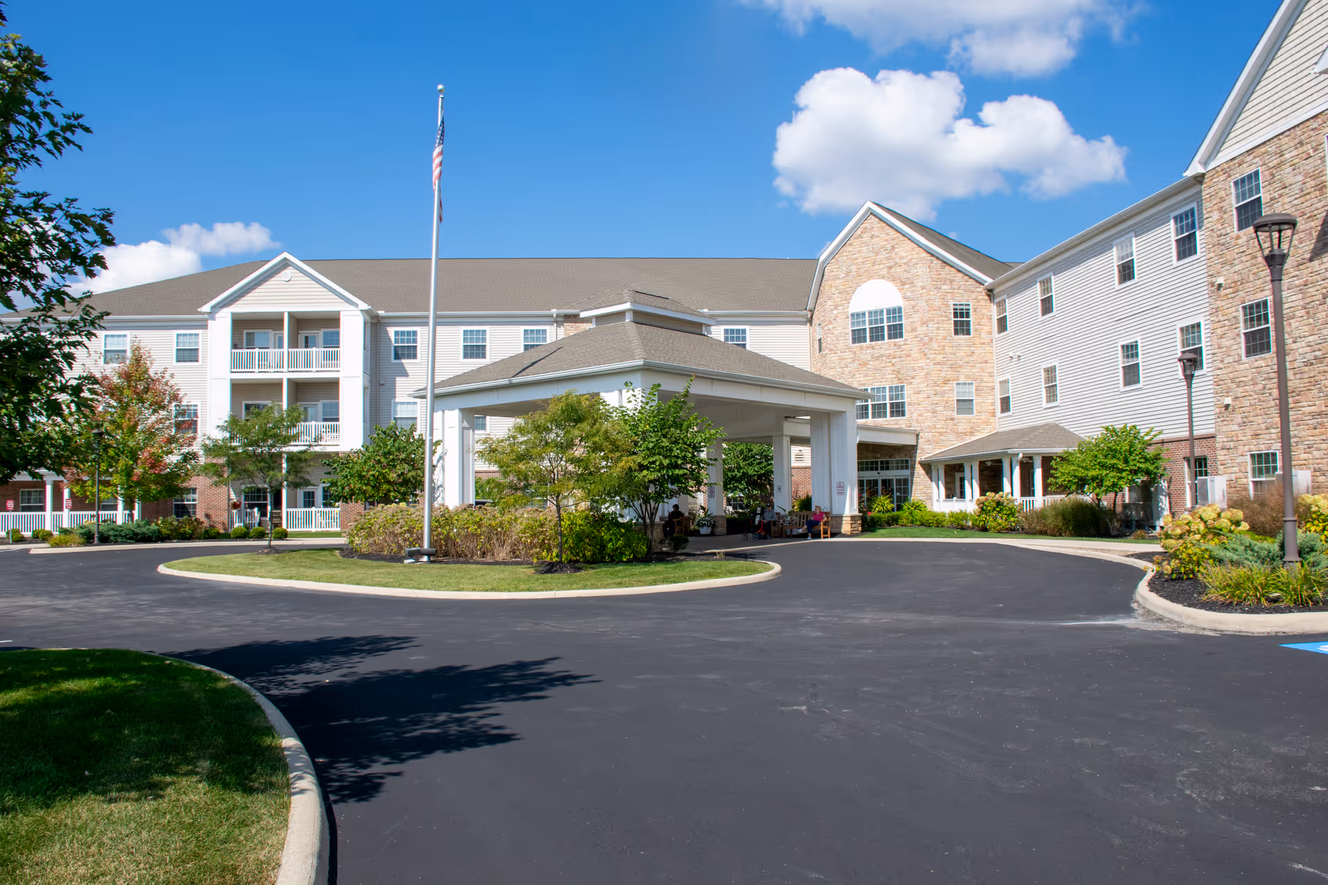 Exterior view of a senior living facility named Danbury Hudson on a sunny day. The building is multi-story with a combination of brick and siding, featuring balconies and large windows. There is a circular driveway with a covered entrance and an American flag on a flagpole in the center. Trees and shrubs are landscaped around the driveway.