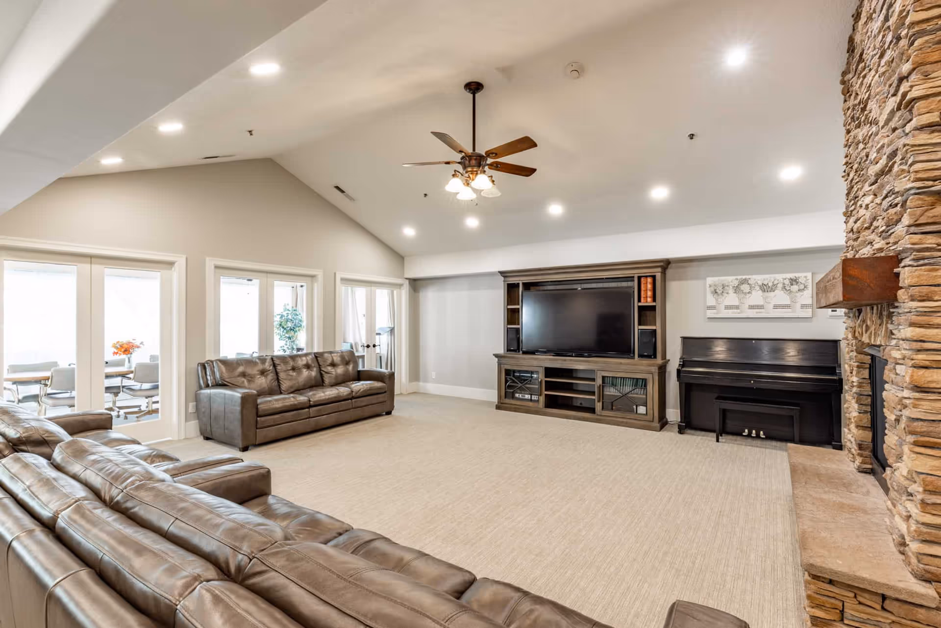 Spacious living room with two brown leather sofas, a large flat-screen TV on a wooden entertainment center, a black piano, and a stone fireplace. The room has a high vaulted ceiling with recessed lighting and a ceiling fan. French doors lead to an adjacent dining area with a table and chairs.