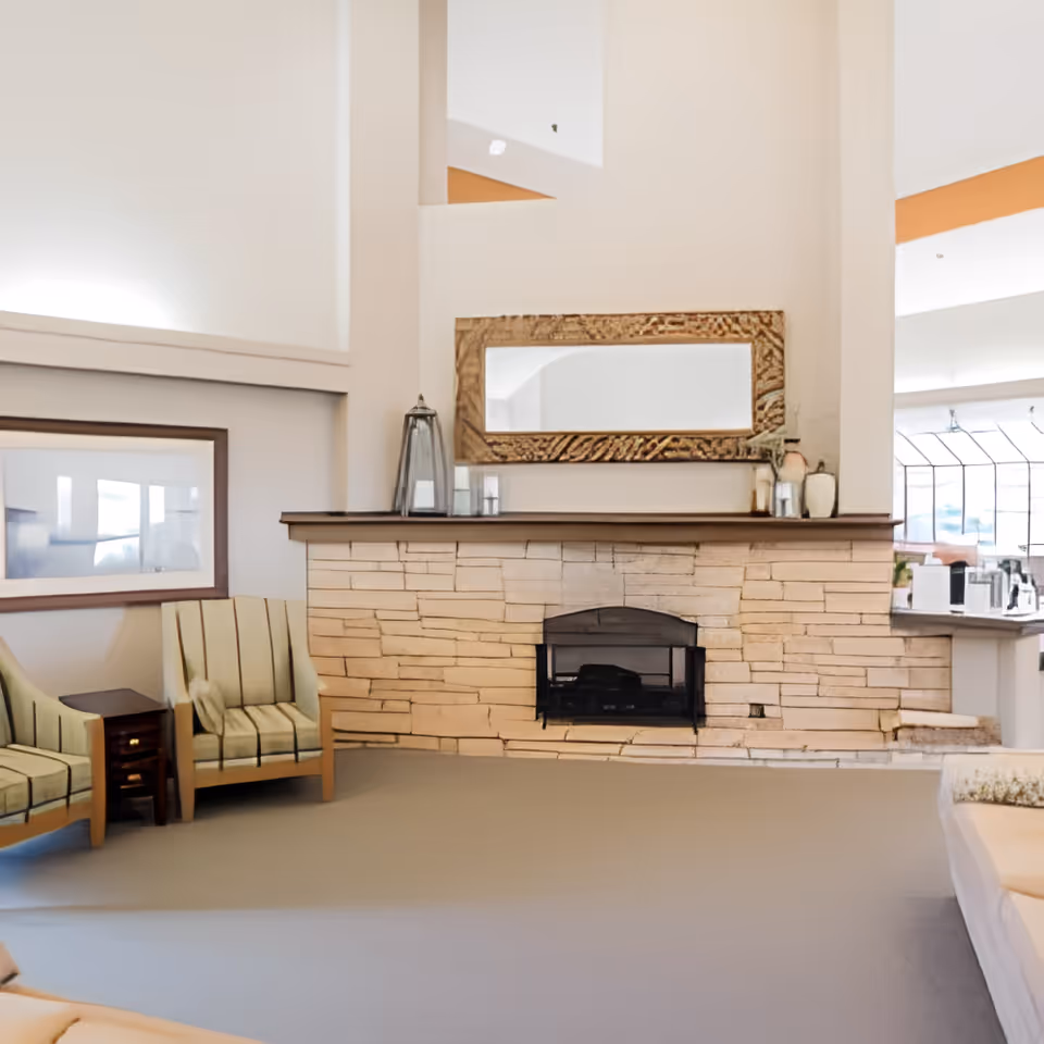 A cozy living room area featuring a stone fireplace with a wooden mantel. Above the mantel is a large rectangular mirror with a textured wooden frame. To the left of the fireplace are two striped armchairs with a small wooden side table between them. The room has high ceilings and light-colored walls, creating a bright and inviting atmosphere.