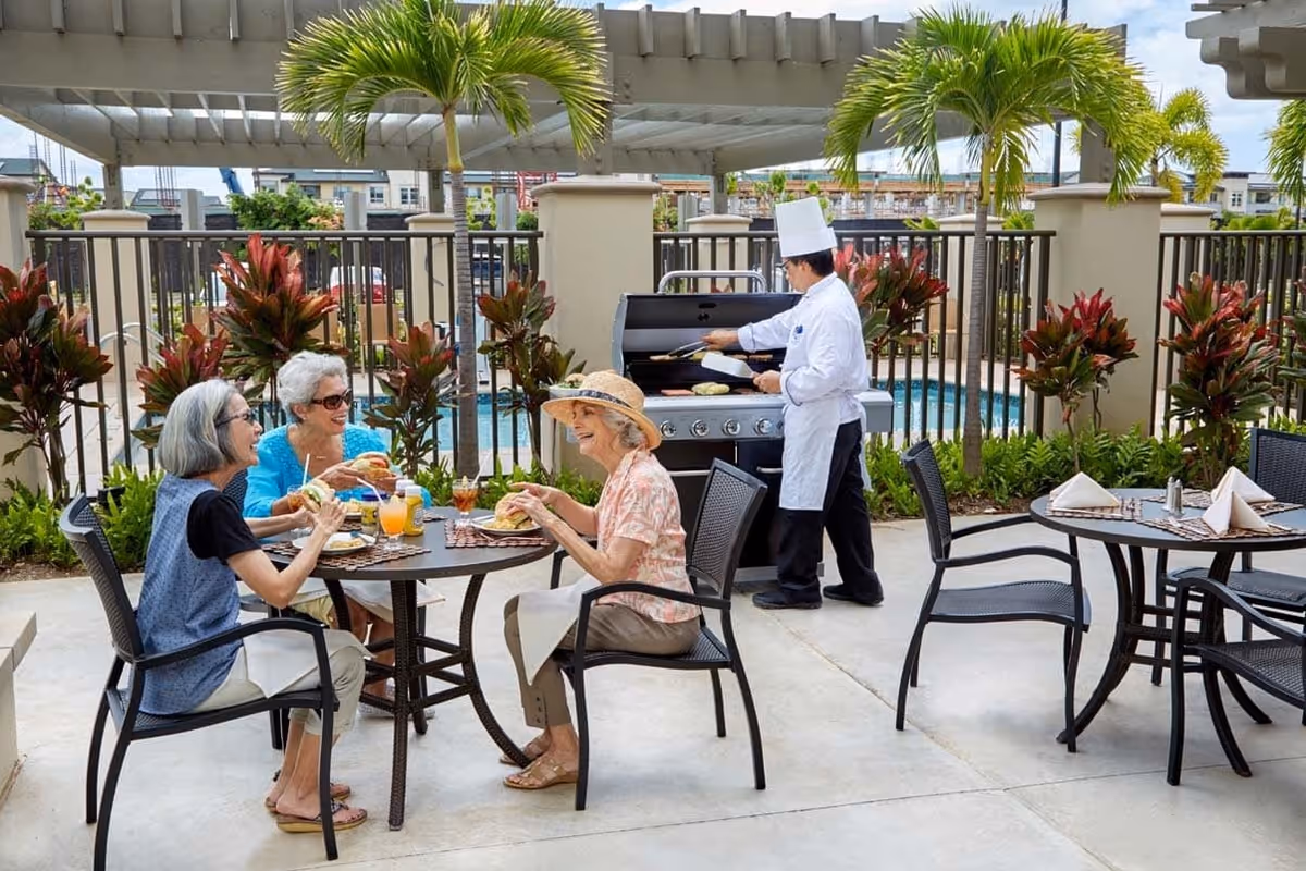 Three older women sit at an outdoor patio table enjoying food and drinks while a chef cooks at a grill near a pool.