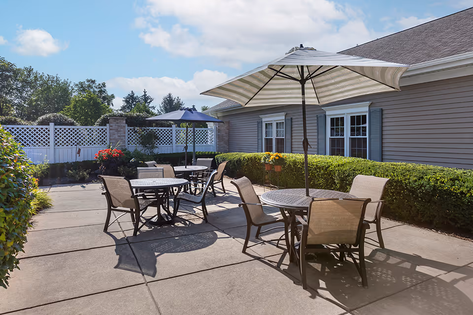 Outdoor patio with round metal tables, chairs, and umbrellas next to a single-story building and trimmed hedges.