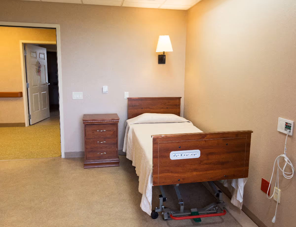 A simple senior facility bedroom with a single adjustable wooden bed, bedside table, and wall lamp.