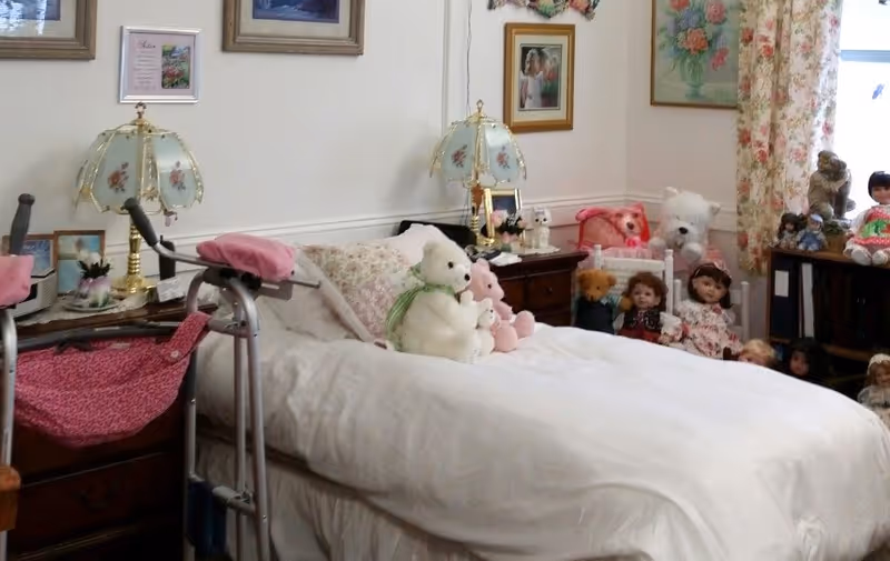 A cozy bedroom with a single bed covered in white bedding and floral pillows. Several stuffed animals and dolls are arranged on the bed and around the room. There are two bedside tables with vintage-style lamps featuring floral designs. The walls are decorated with framed pictures and floral curtains hang by the window.