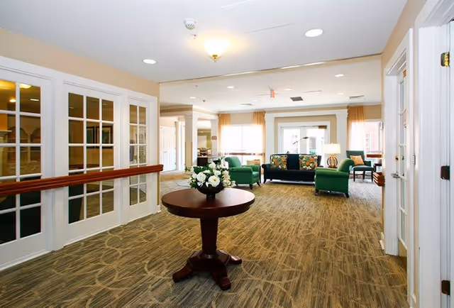 A bright and spacious senior living community common area with beige walls and carpeted floor. In the foreground, there is a round wooden table with a floral arrangement. In the background, there are green upholstered chairs and a dark sofa with colorful cushions near large windows letting in natural light.