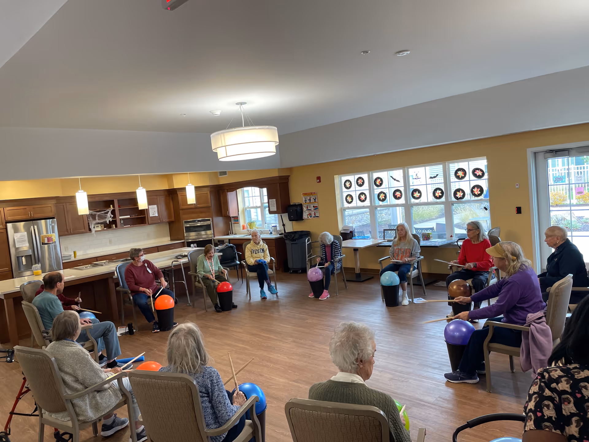 A circle of older adults seated in a senior living community room participating in a seated exercise activity with colorful exercise balls.