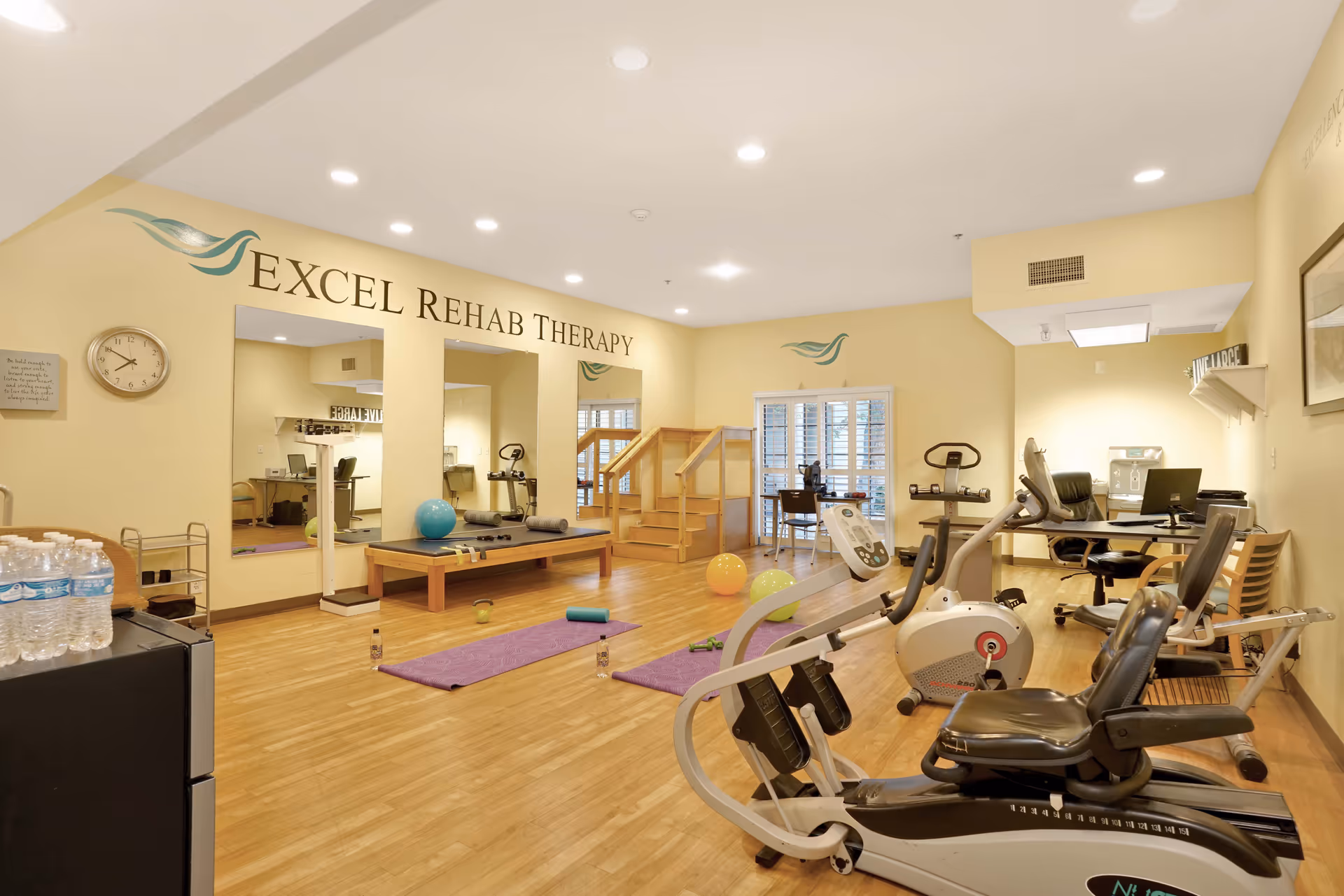 Bright rehab therapy room with exercise machines, therapy mats, stability balls and a treatment table beneath a wall sign reading 'Excel Rehab Therapy'.