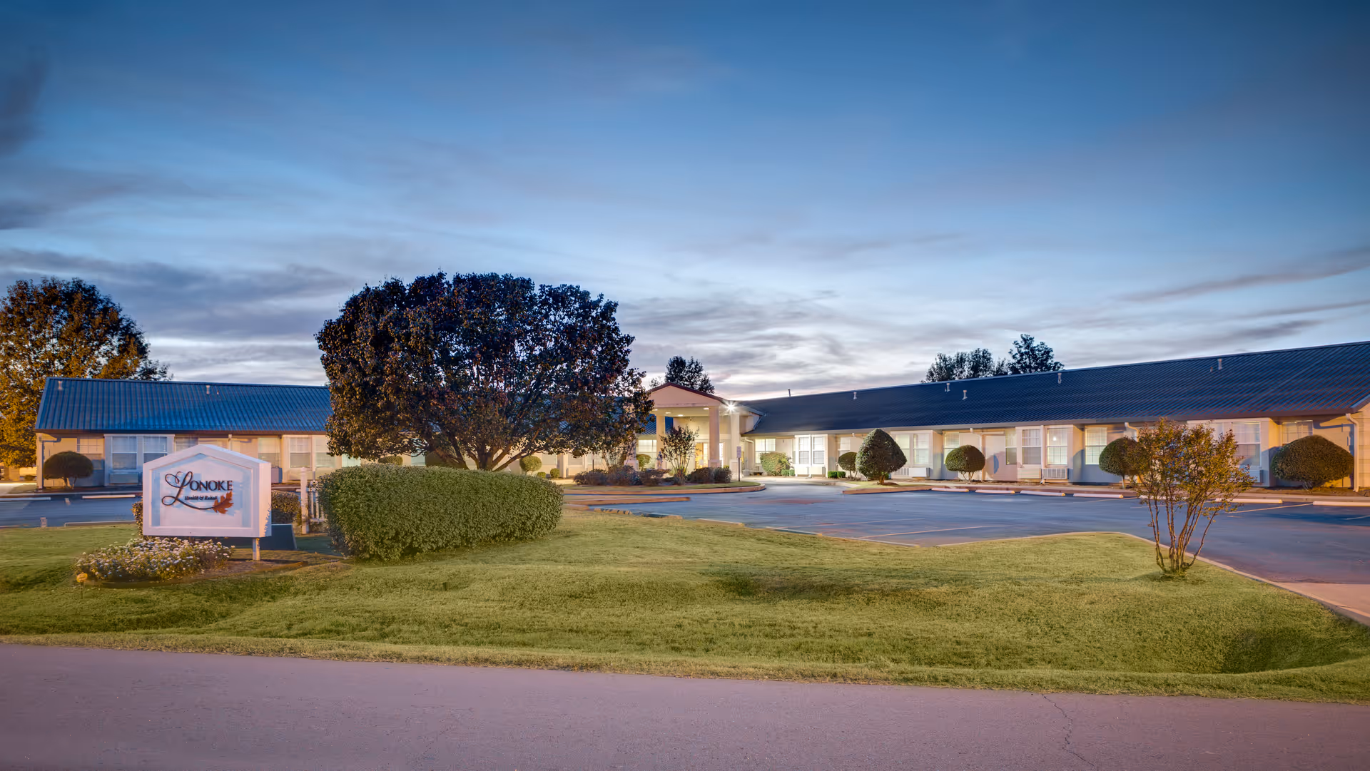 Single-story Lonoke Nursing & Rehab Center building and sign on a landscaped lawn at dusk.