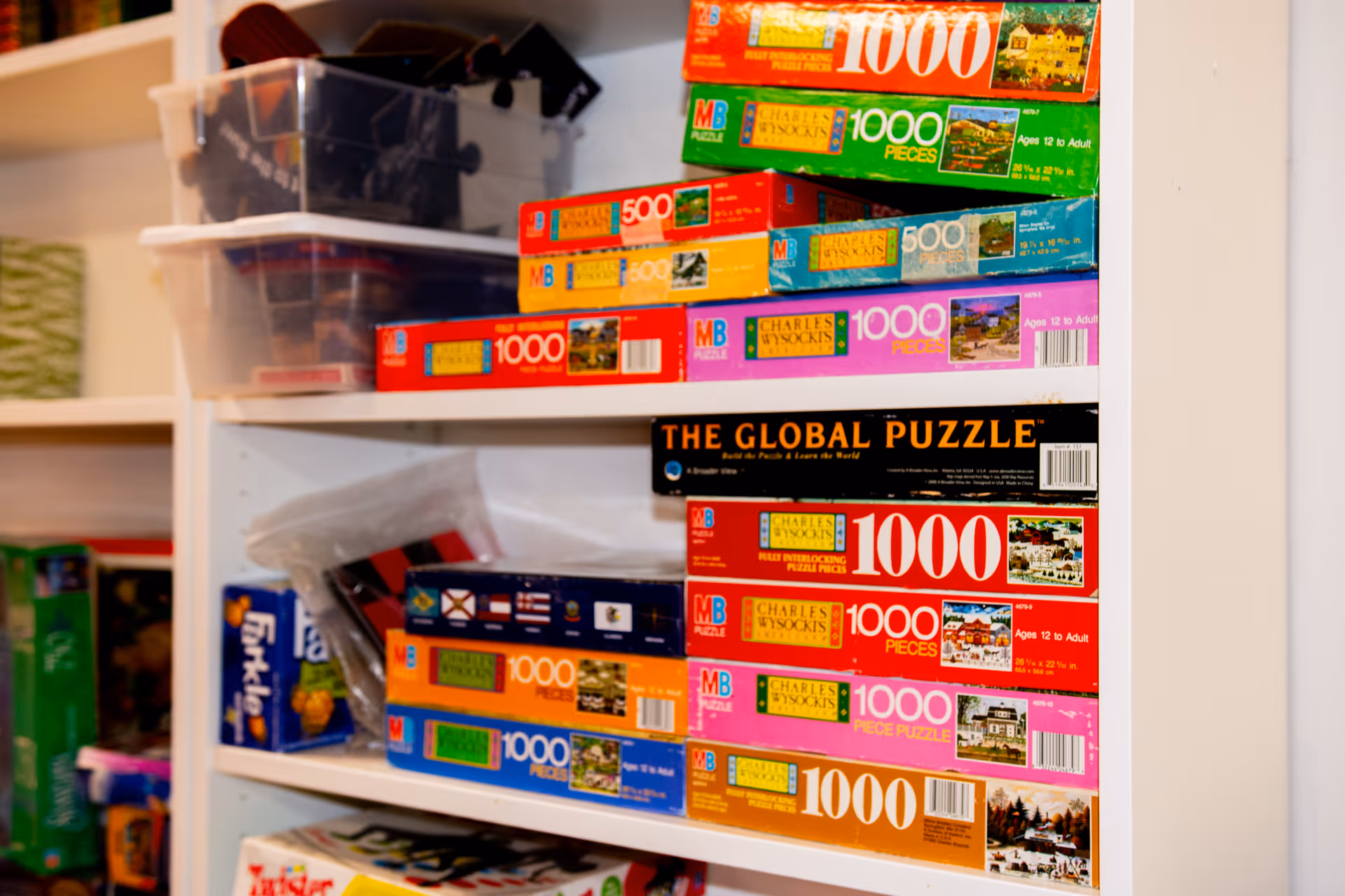 Shelves filled with various boxed puzzles, including multiple 1000-piece puzzles by Charles Wysocki and a puzzle titled 'The Global Puzzle'. There are also clear plastic storage containers on the top shelf and other board games visible on the lower shelves.