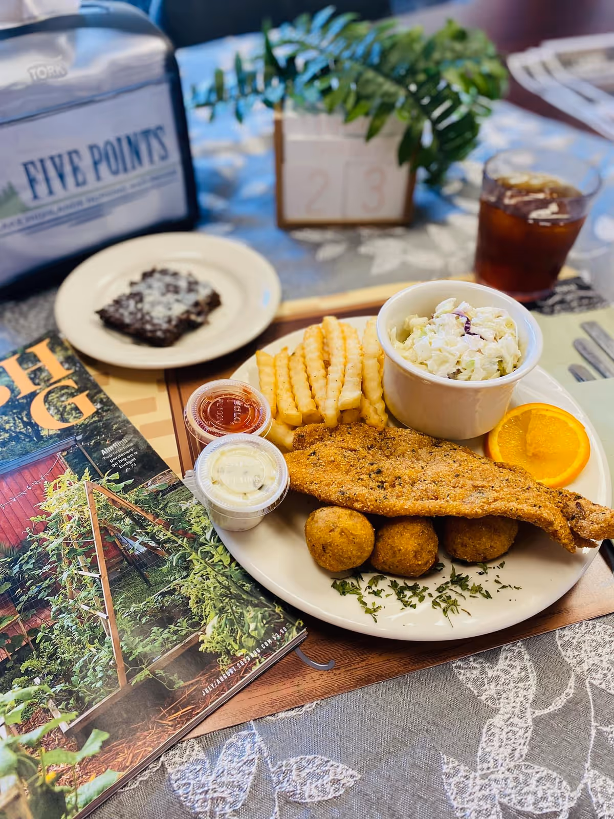A plate of food on a table featuring fried fish, crinkle-cut fries, hush puppies, coleslaw, and a slice of orange. Two small containers of dipping sauces are on the plate. Nearby is a plate with a brownie, a glass of iced tea, a napkin holder with the text 'FIVE POINTS', a table number block showing '23', and a magazine with a garden on the cover. The tablecloth has a floral pattern.
