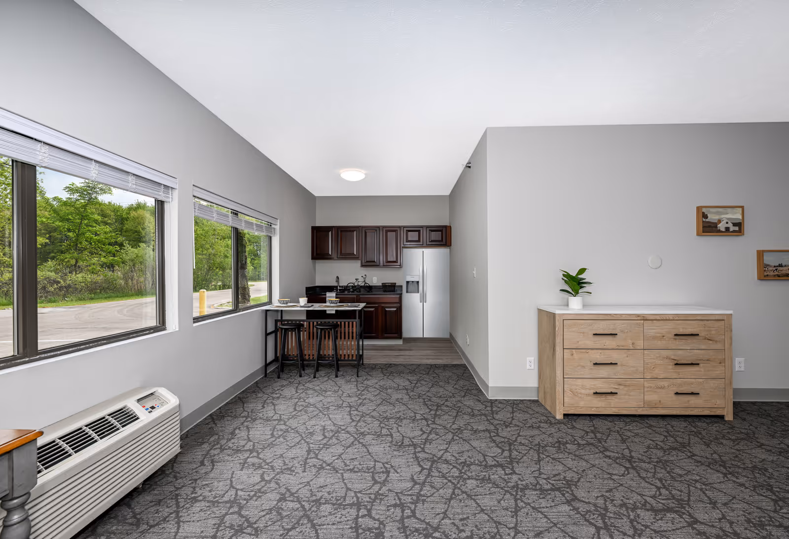 Interior view of a senior living facility room with large windows on the left side showing greenery outside. The room features a small kitchen area with dark wood cabinets, a stainless steel refrigerator, and a small dining table with three stools. On the right side, there is a wooden dresser with a small potted plant on top and two framed pictures hanging on the wall above it. The floor is carpeted with a gray patterned design.