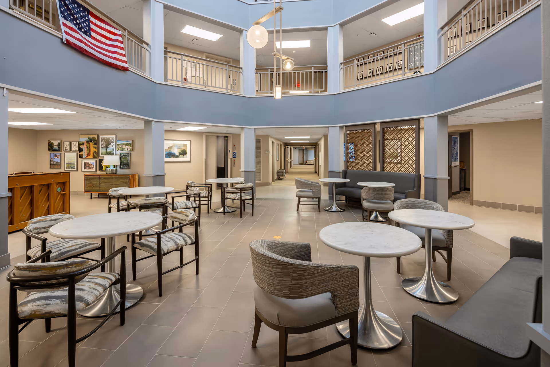 A spacious, modern common area in a senior living facility with multiple round tables and cushioned chairs arranged for seating. The area features a two-story ceiling with a balcony railing on the upper floor, a large American flag hanging on the left side, and various framed pictures on the walls. The lighting is bright with ceiling lights and a hanging pendant light fixture.