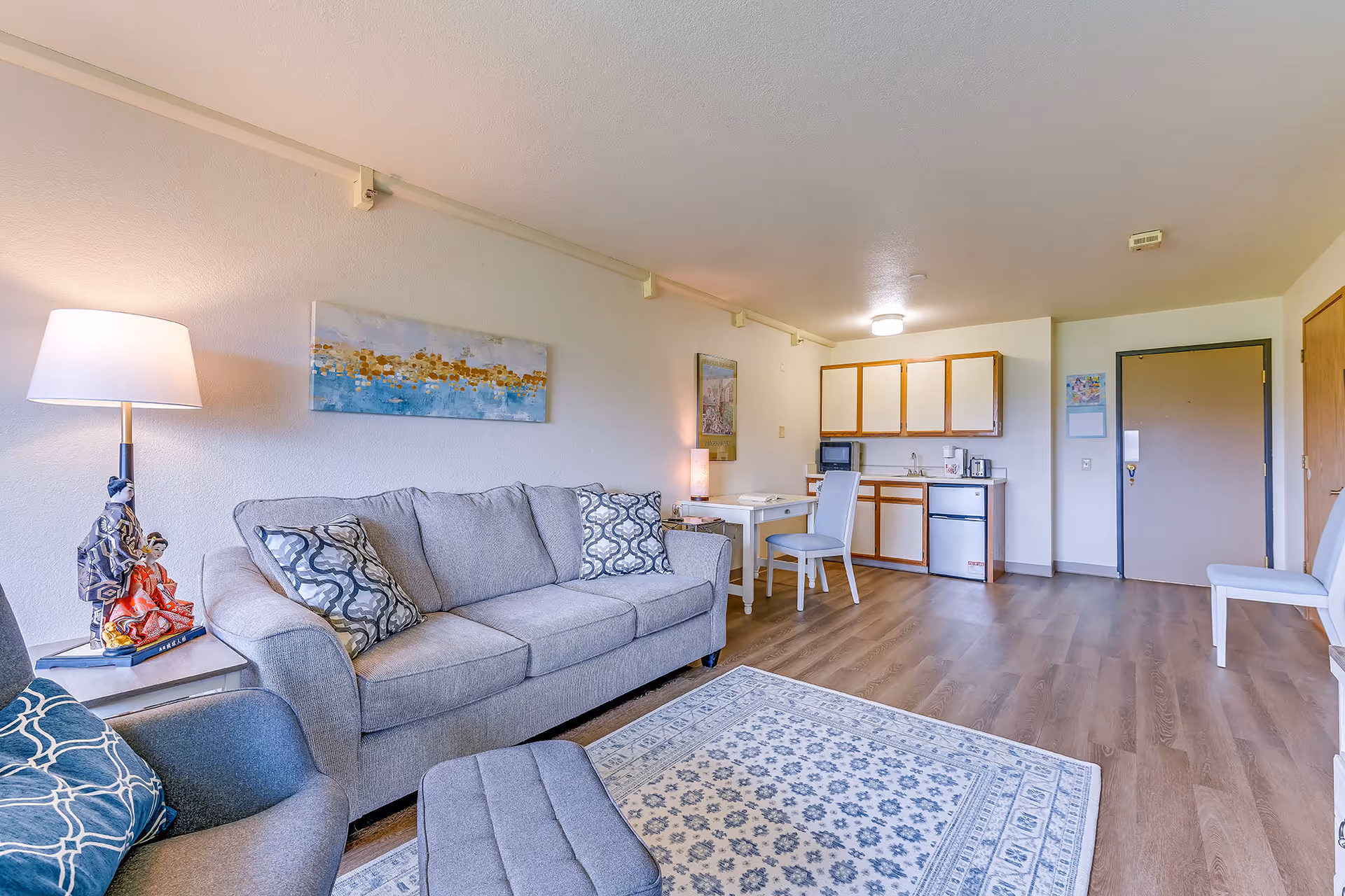 A bright and clean senior living apartment interior featuring a gray sofa with patterned pillows, a matching armchair with a blue pillow, a small ottoman, a floor lamp, and decorative figurines on a side table. The room has light wood flooring and a patterned area rug. In the background, there is a kitchenette with white cabinets, a small refrigerator, microwave, coffee maker, and a small dining table with two chairs. The entrance door and a closet door are visible on the right side of the room.