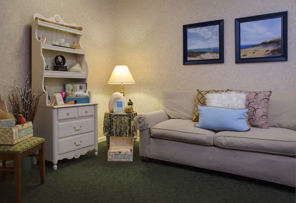 Cozy living room area with a beige sofa adorned with multiple decorative pillows, a small round table covered with a patterned cloth holding a lamp and decorative items, a white wooden cabinet with shelves displaying various ornaments, and two framed paintings of beach scenes hanging on the wall.