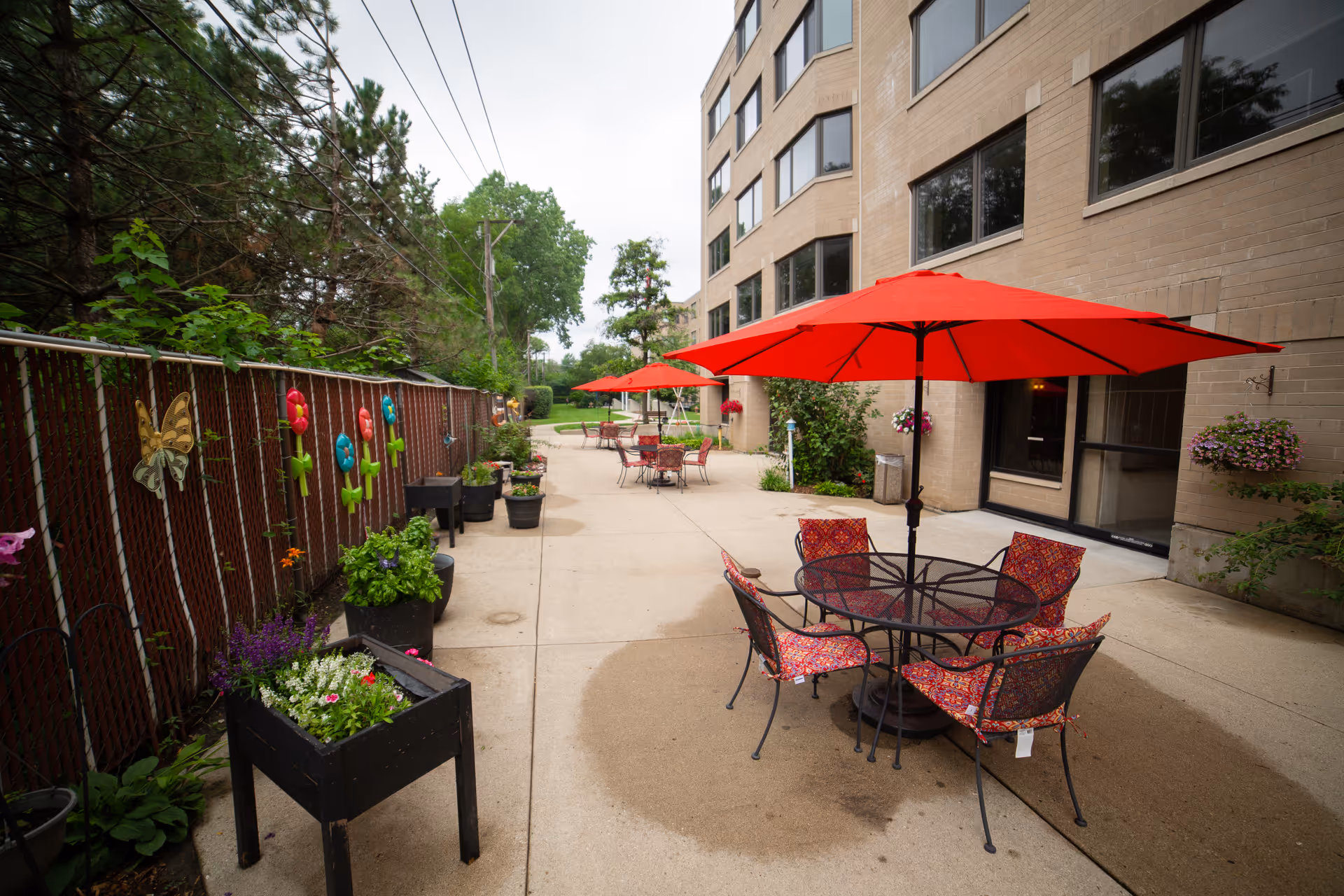 Outdoor patio area at a senior living facility with several round tables and chairs, each shaded by large red umbrellas. The patio is adjacent to a multi-story beige brick building. Along the left side, there is a chain-link fence decorated with colorful flower and butterfly ornaments, and several potted plants are placed along the fence.