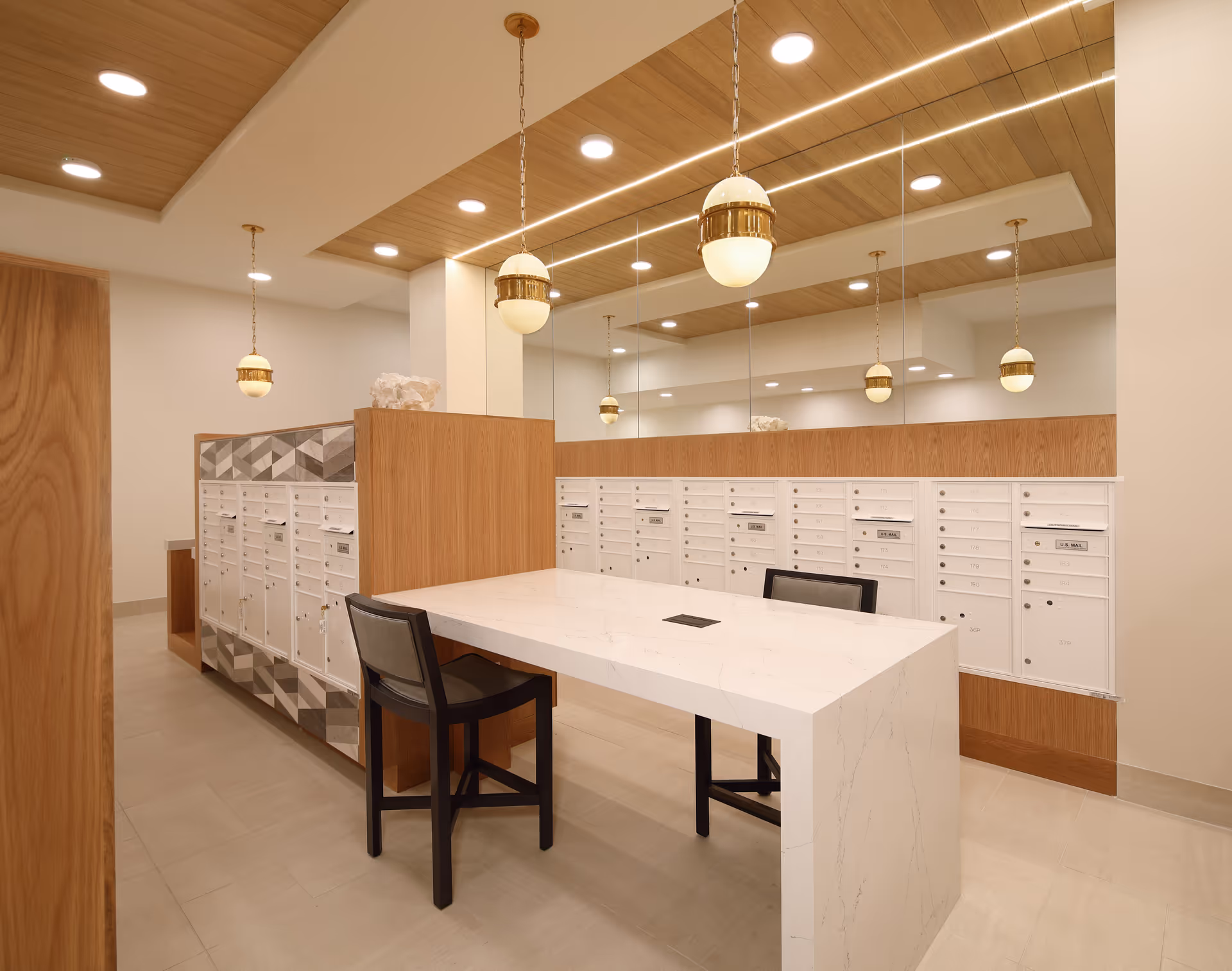 Interior view of a mailroom with multiple white mailboxes mounted on wooden panels. A white marble table with two black chairs is placed in the center. The ceiling features wooden panels with recessed lighting and hanging pendant lights with gold accents. A large mirror covers one wall, reflecting the mailboxes and lights.
