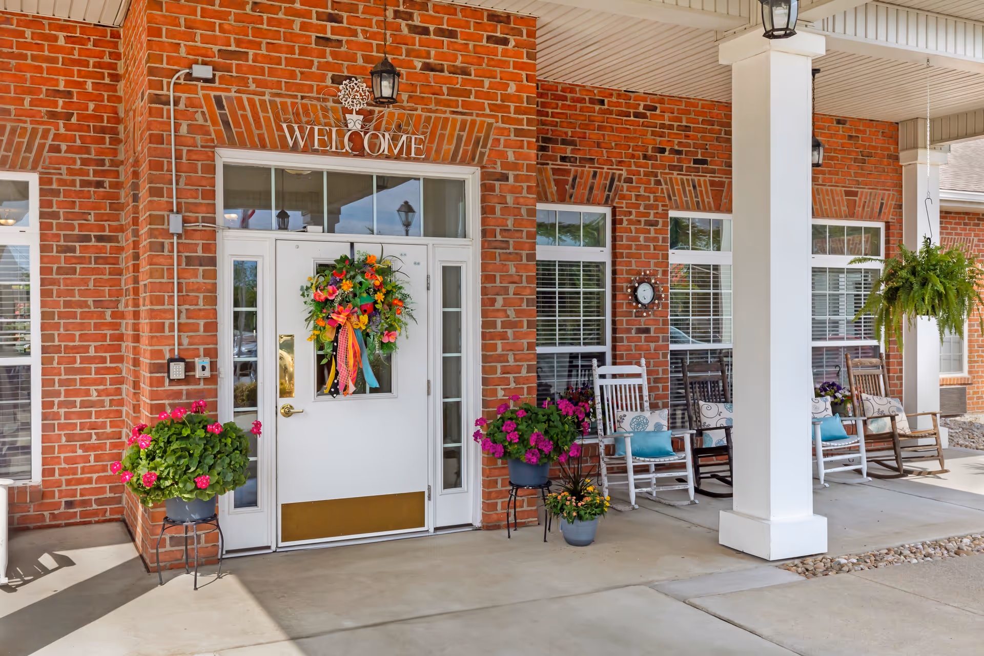 Covered brick entrance with a white door decorated with a colorful wreath, potted flowers, rocking chairs and a 'Welcome' sign.