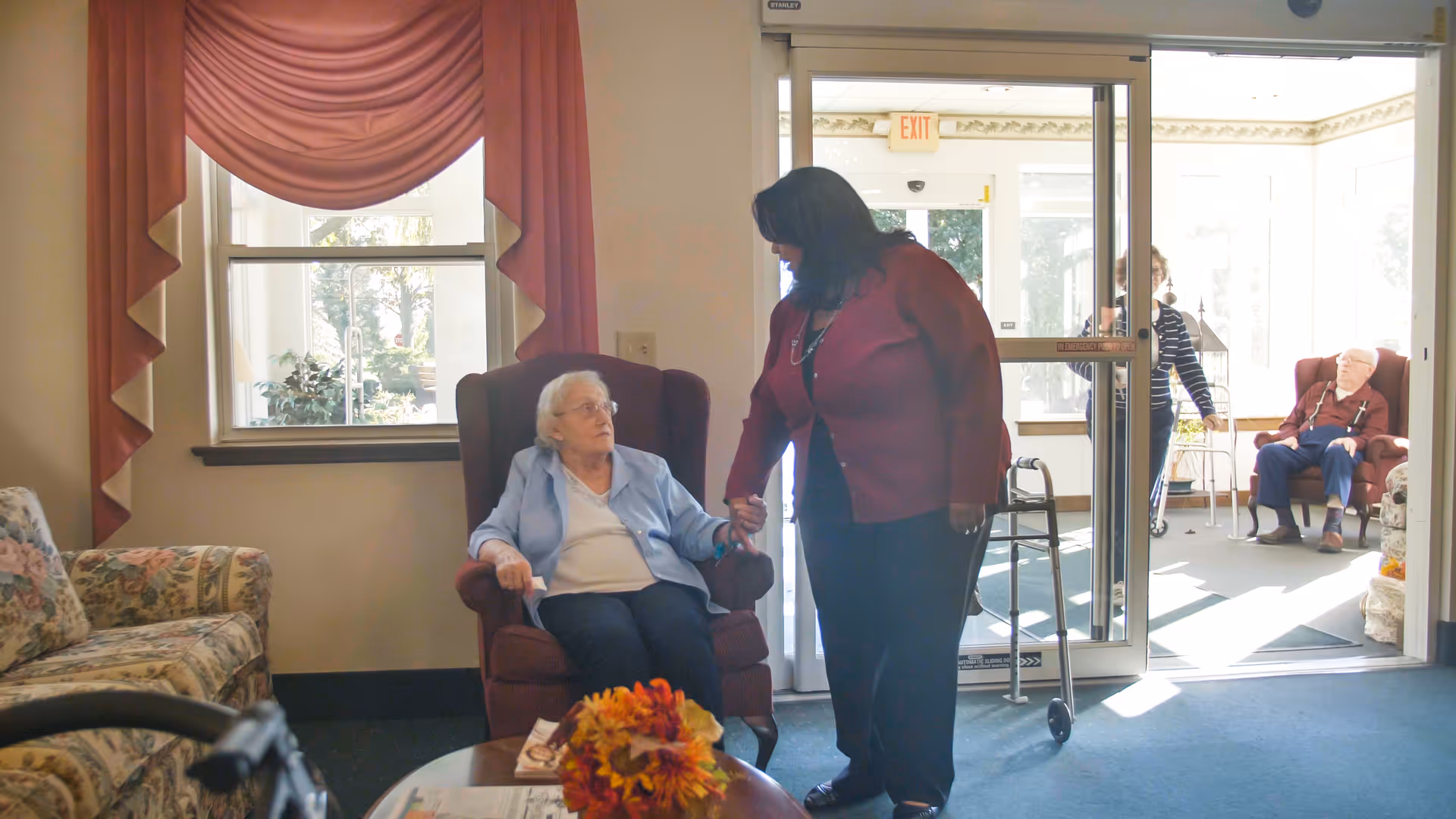 An elderly woman sitting in a maroon armchair holding hands with a standing caregiver in a red jacket inside a well-lit common area. Another elderly woman with a walker and an elderly man sitting in a similar armchair are visible near the glass entrance door. The room has floral patterned furniture and a window with red drapes.