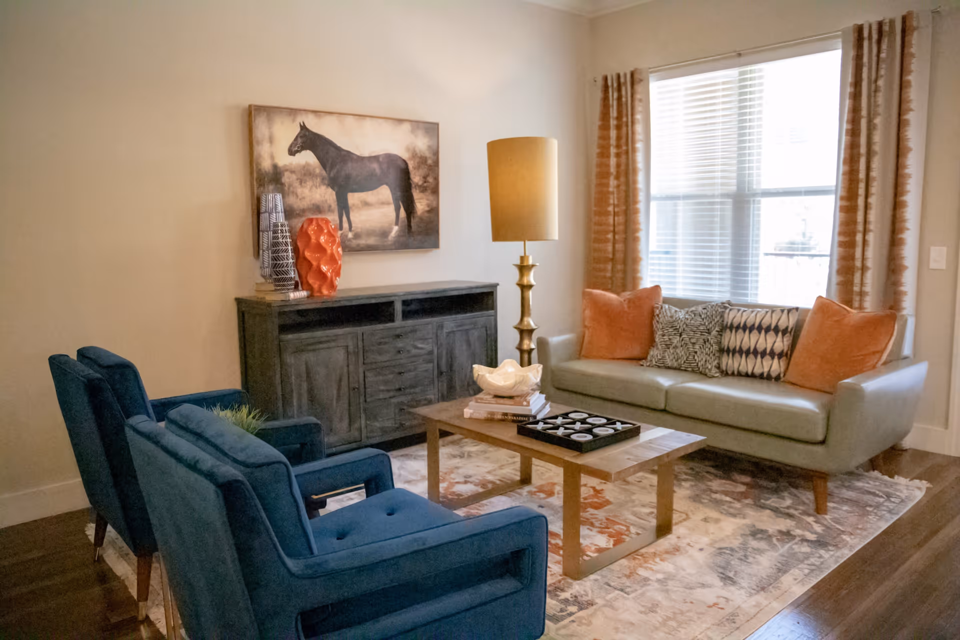 A cozy living room with a light gray sofa adorned with orange and patterned cushions, two blue armchairs, a wooden coffee table with books and a decorative tray, a tall gold floor lamp, a dark wooden cabinet with decorative vases, and a large window with blinds and orange curtains letting in natural light. A painting of a horse hangs on the wall above the cabinet.