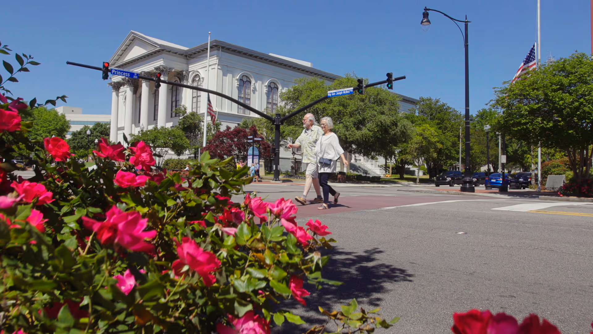 An elderly couple walking across a street intersection on a sunny day with bright pink flowers in the foreground and a large white classical building with columns in the background. The street signs read Princess St and N. Orange St.