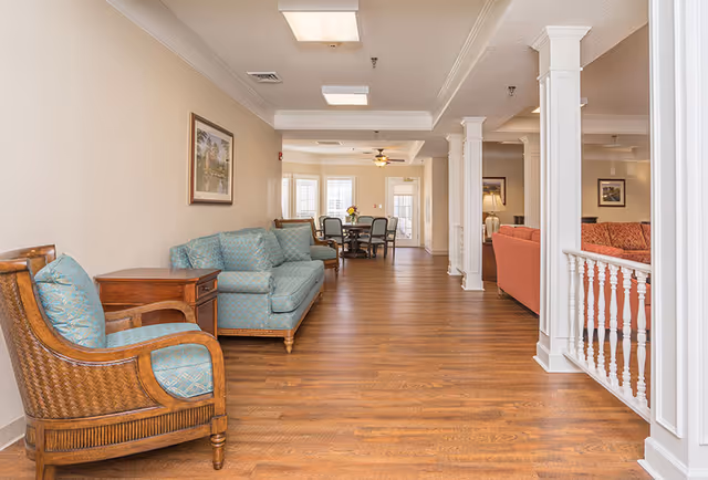 A spacious senior living facility common area with wooden flooring, light beige walls, and white ceiling. The room features a blue cushioned armchair and sofa on the left, a wooden side table, and a dining table with chairs in the background near windows with white blinds. White columns and a white railing separate the seating areas, and framed artwork hangs on the walls.